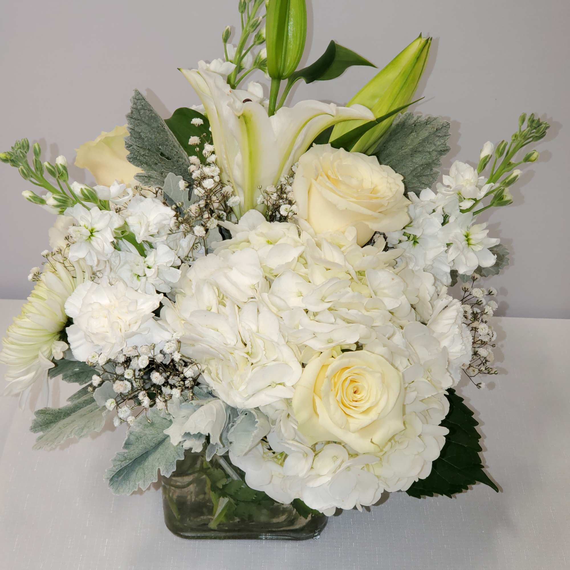 Low white and cream flower arrangement with lilies, roses, and hydrangeas in a glass cube vase