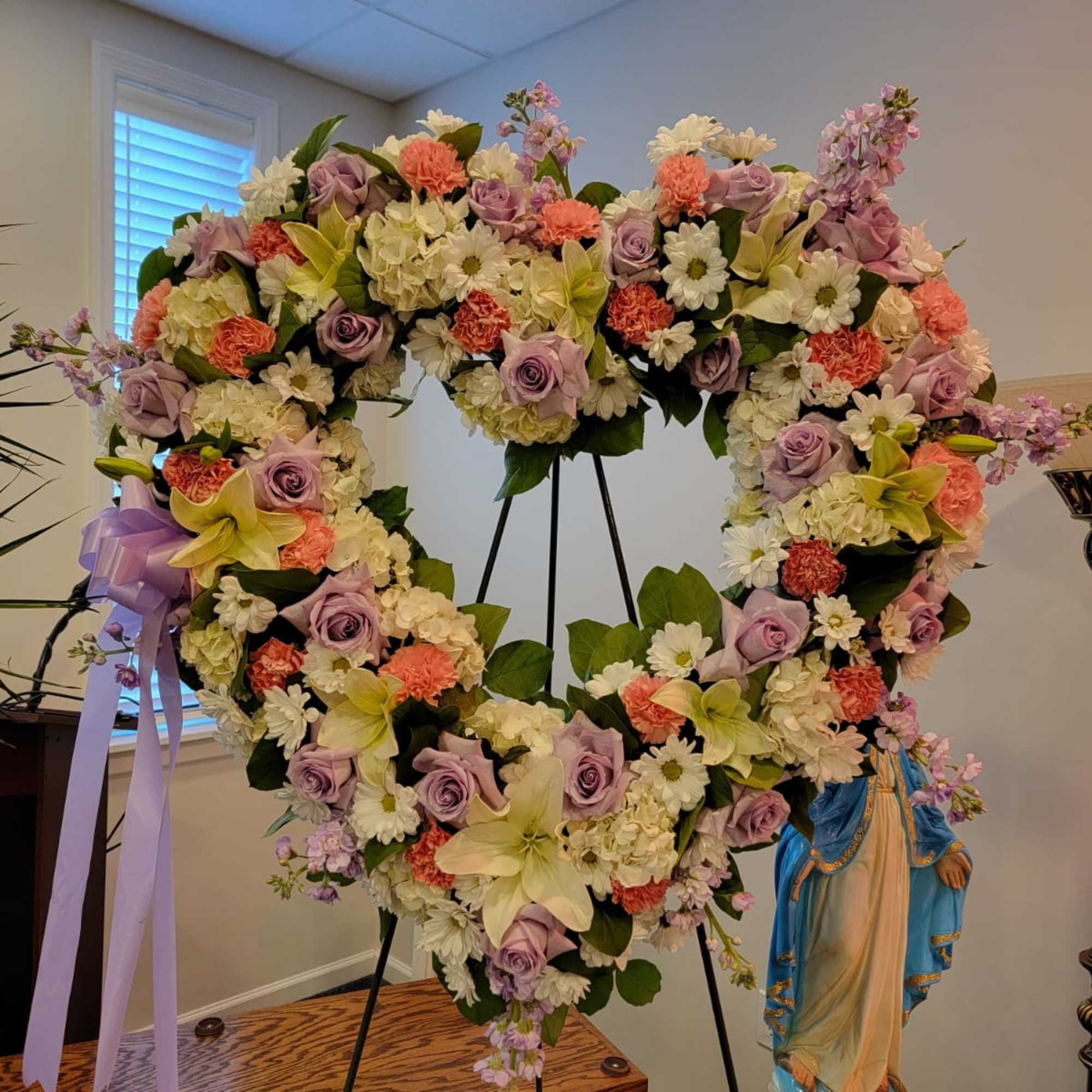 Heart-shaped standing wreath of lavender roses, white lilies, and pastel flowers on a metal easel.