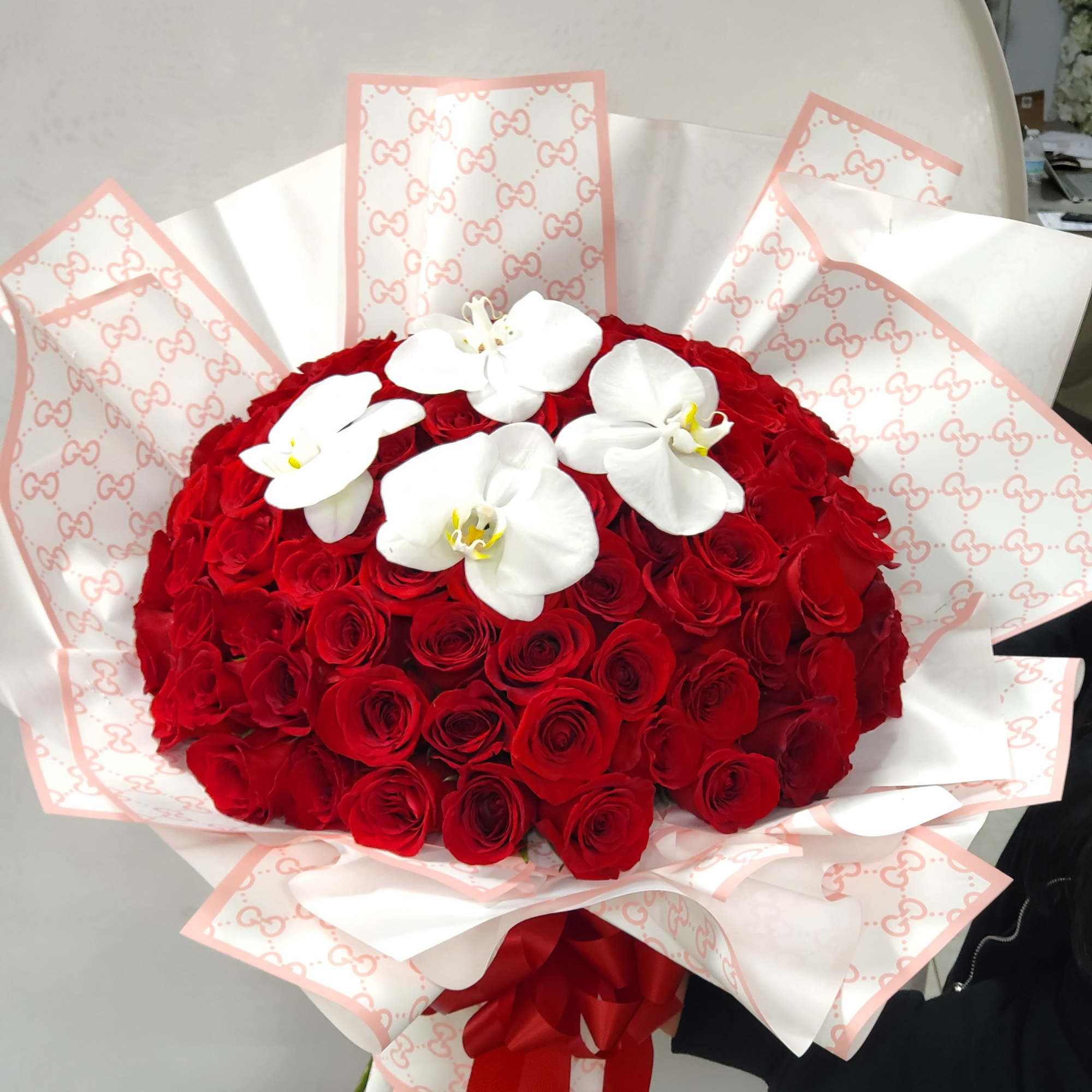 Large bouquet of tightly packed red roses topped with white orchids, wrapped in patterned white paper with red ribbon