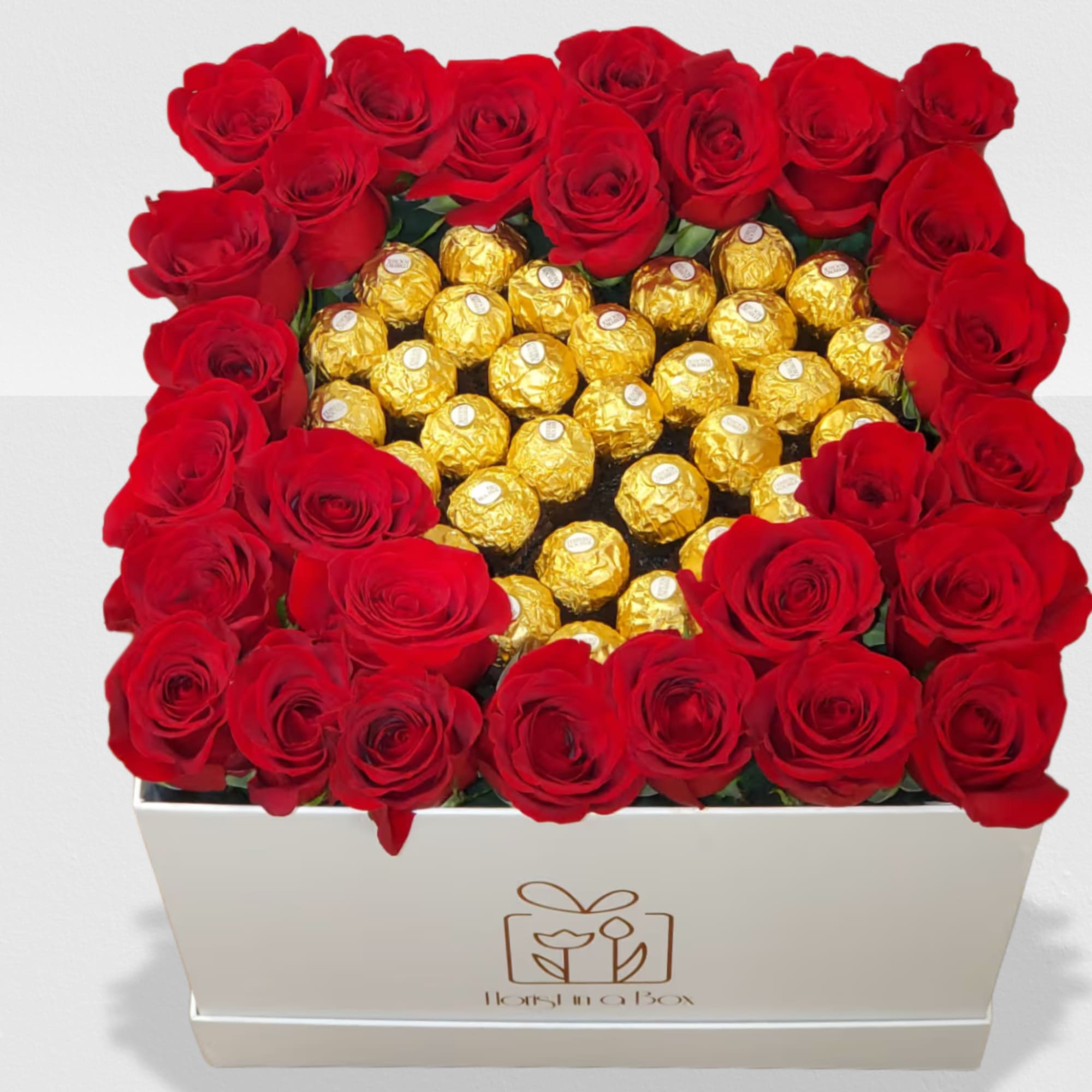 Square white box filled with red roses framing gold-wrapped chocolates in the center