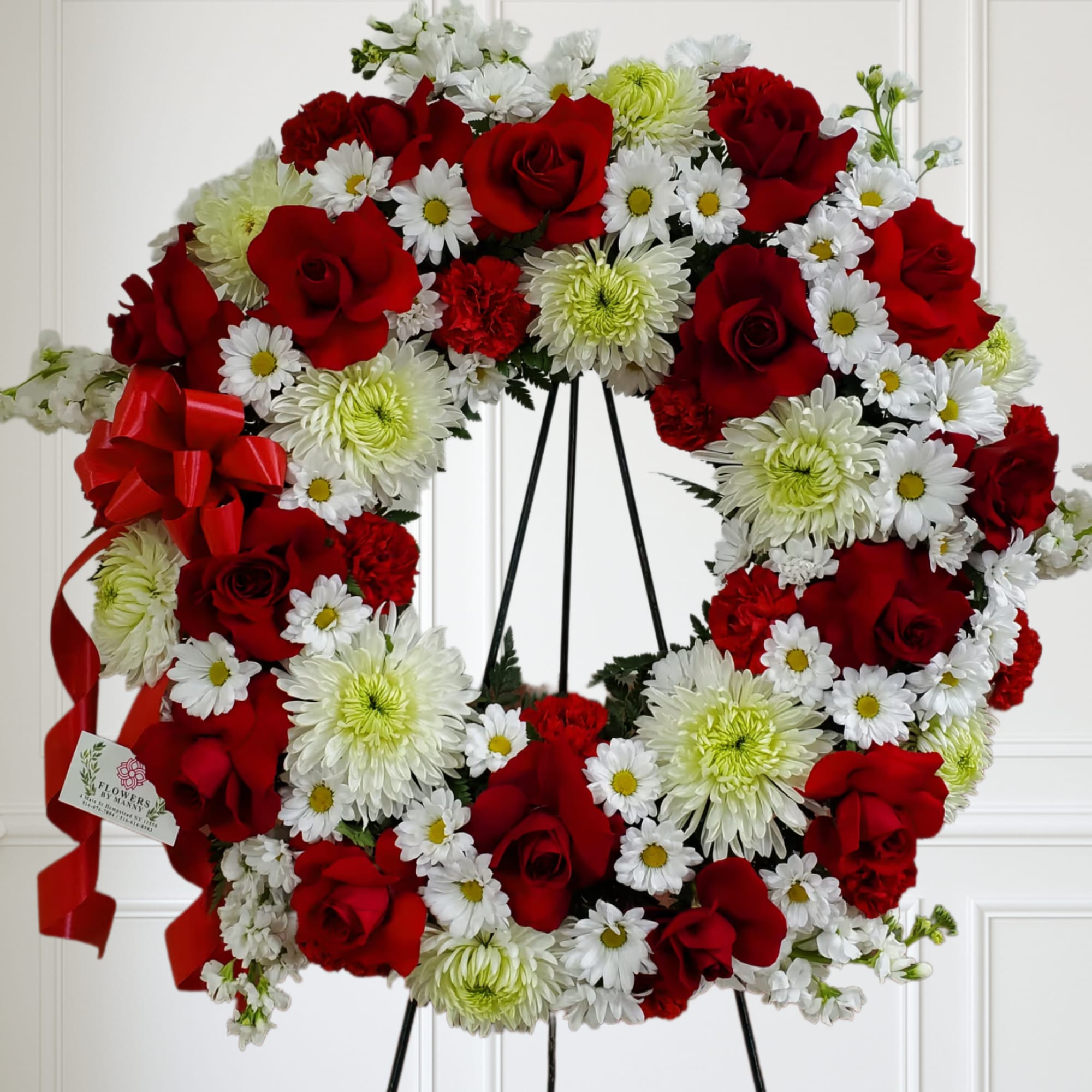 Standing wreath of red roses, white daisies, and green-tinted mums on a black easel with red ribbon bow