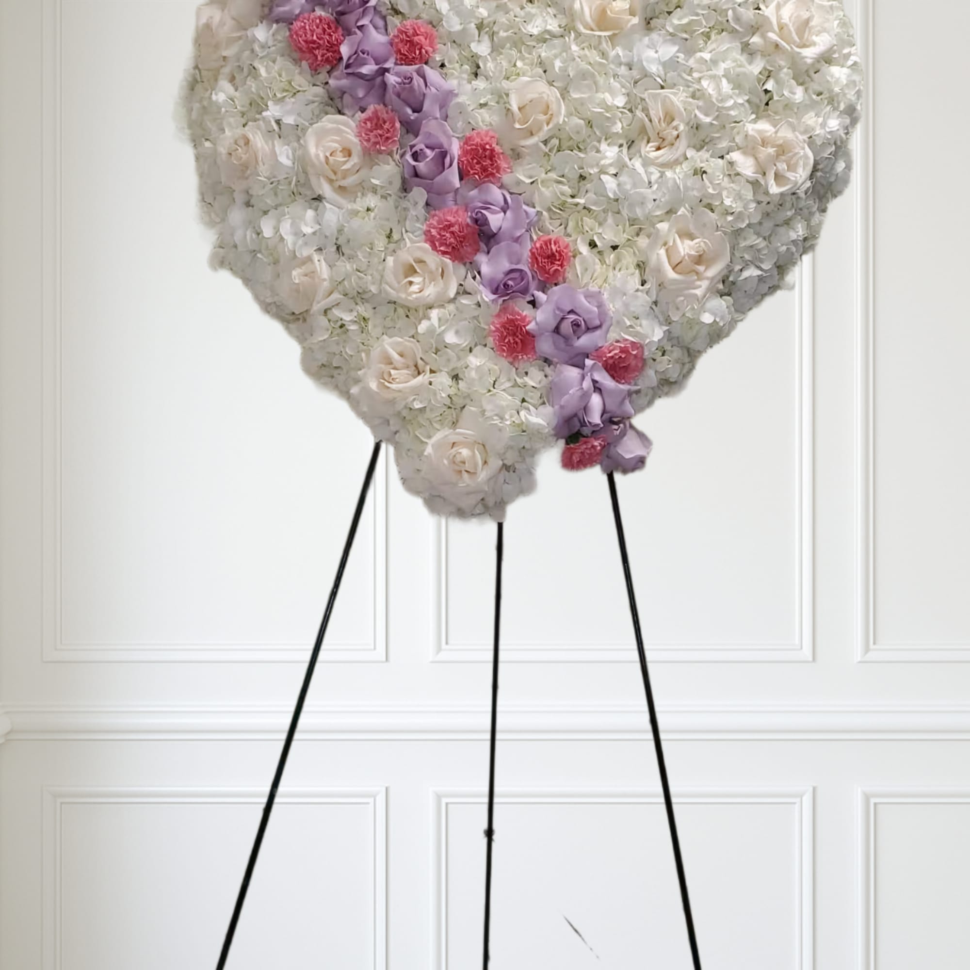 Heart-shaped standing spray of white flowers with cream roses, lavender blooms, and pink carnations on a black easel
