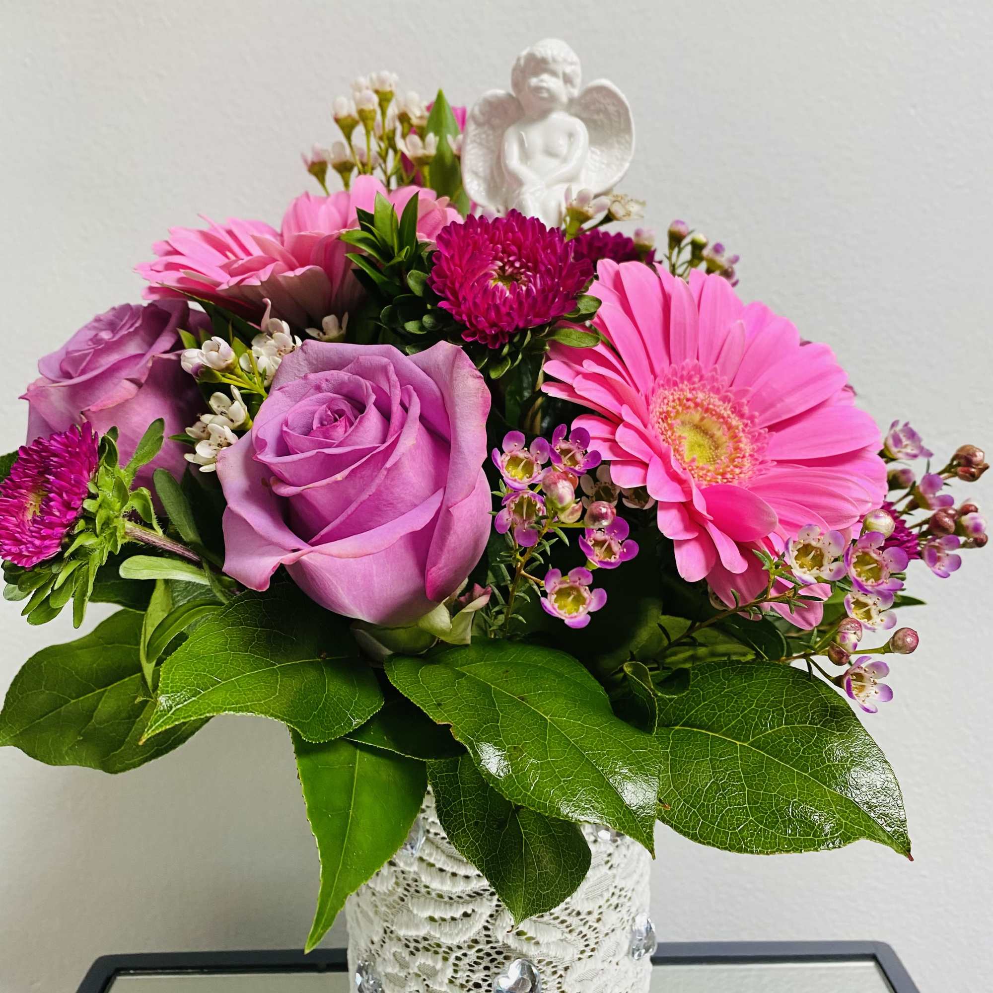 Pink roses and gerbera daisies in a lace-wrapped vase with a small angel figurine