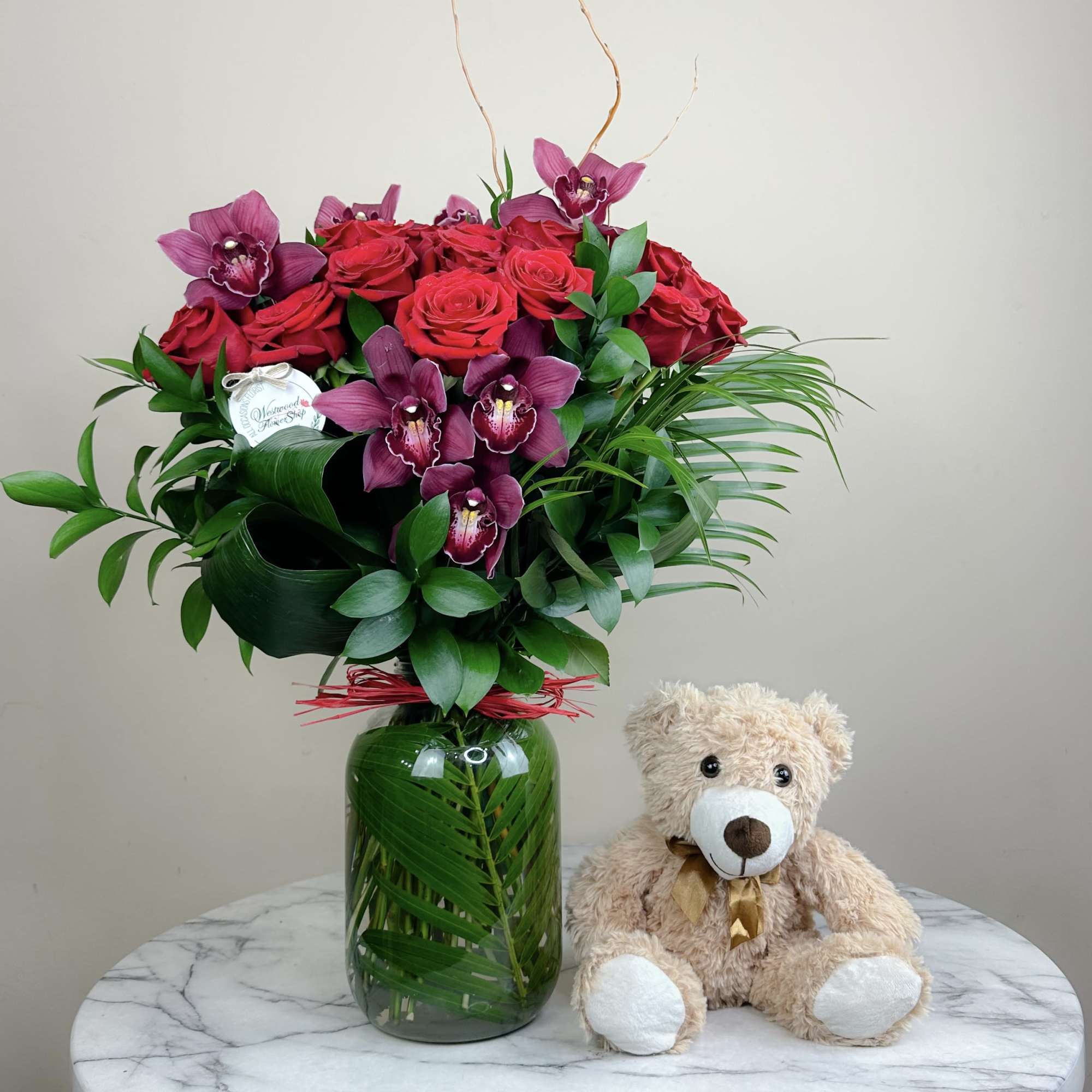 Red roses and purple orchids in a glass vase beside a teddy bear