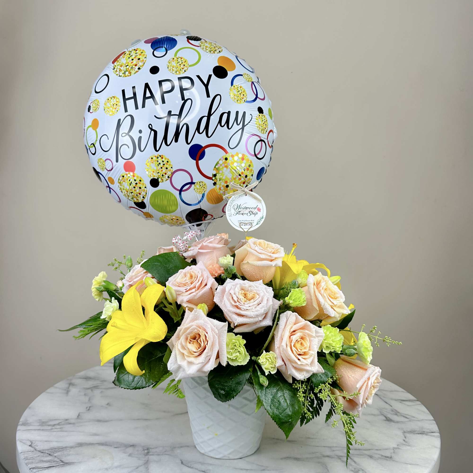 Birthday balloon above a bouquet of pale roses and yellow lilies in a white vase