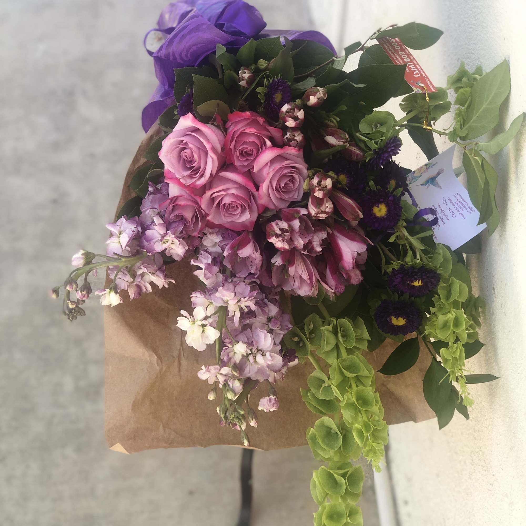 Bouquet of pink roses, purple flowers, and green hanging blooms in brown paper