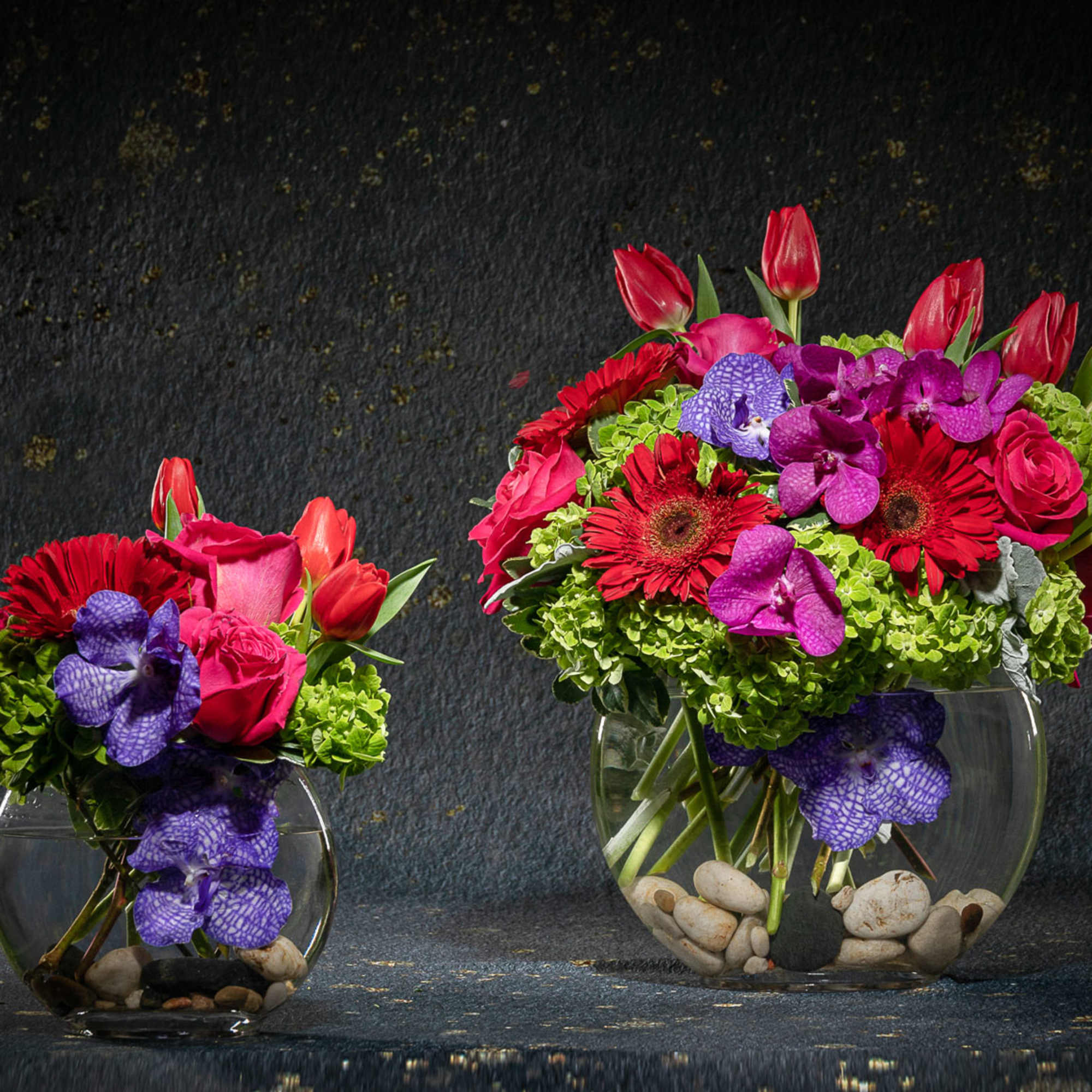 Two colorful flower arrangements in round glass vases on a dark background.