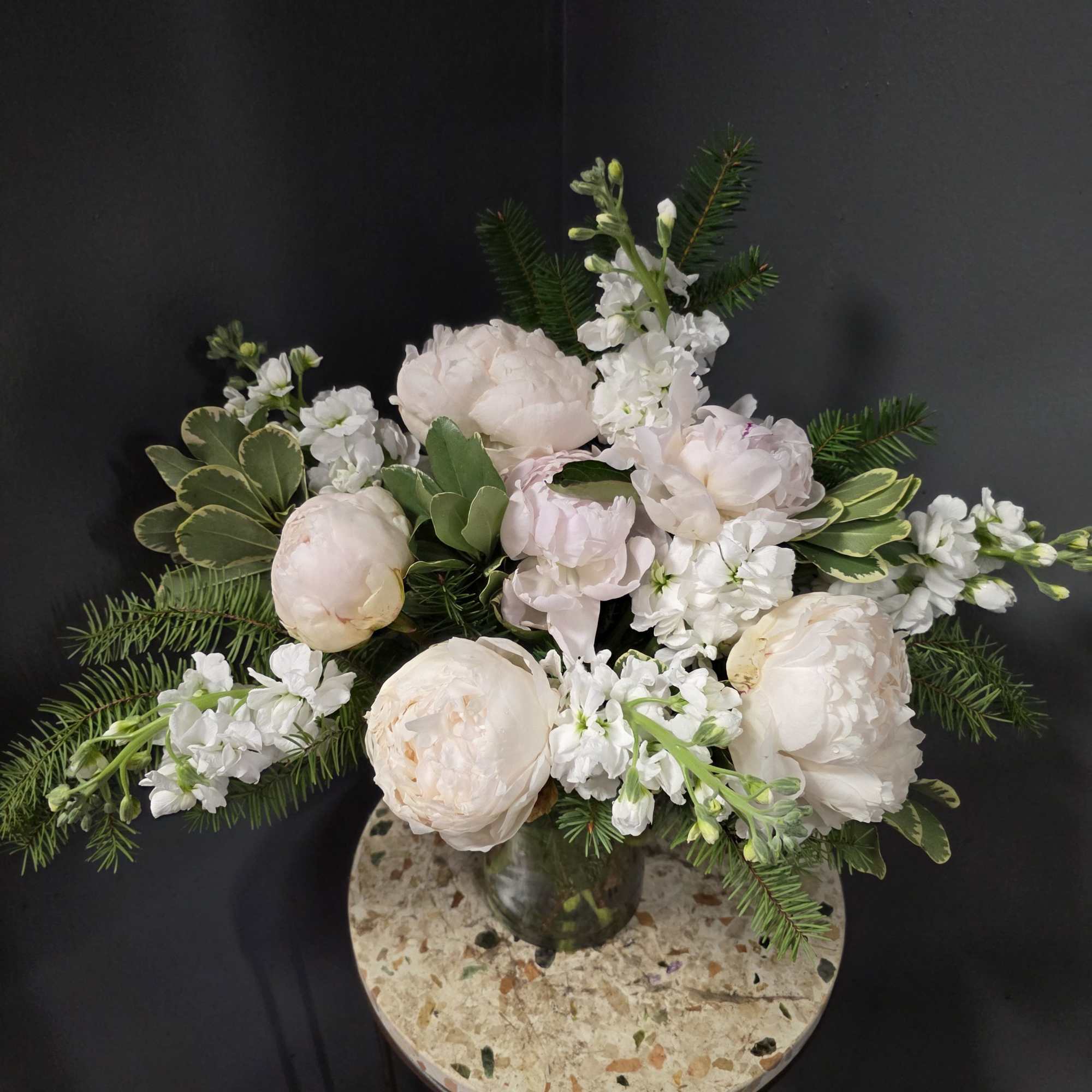 Soft pink peonies and white flowers in a glass vase with evergreen foliage