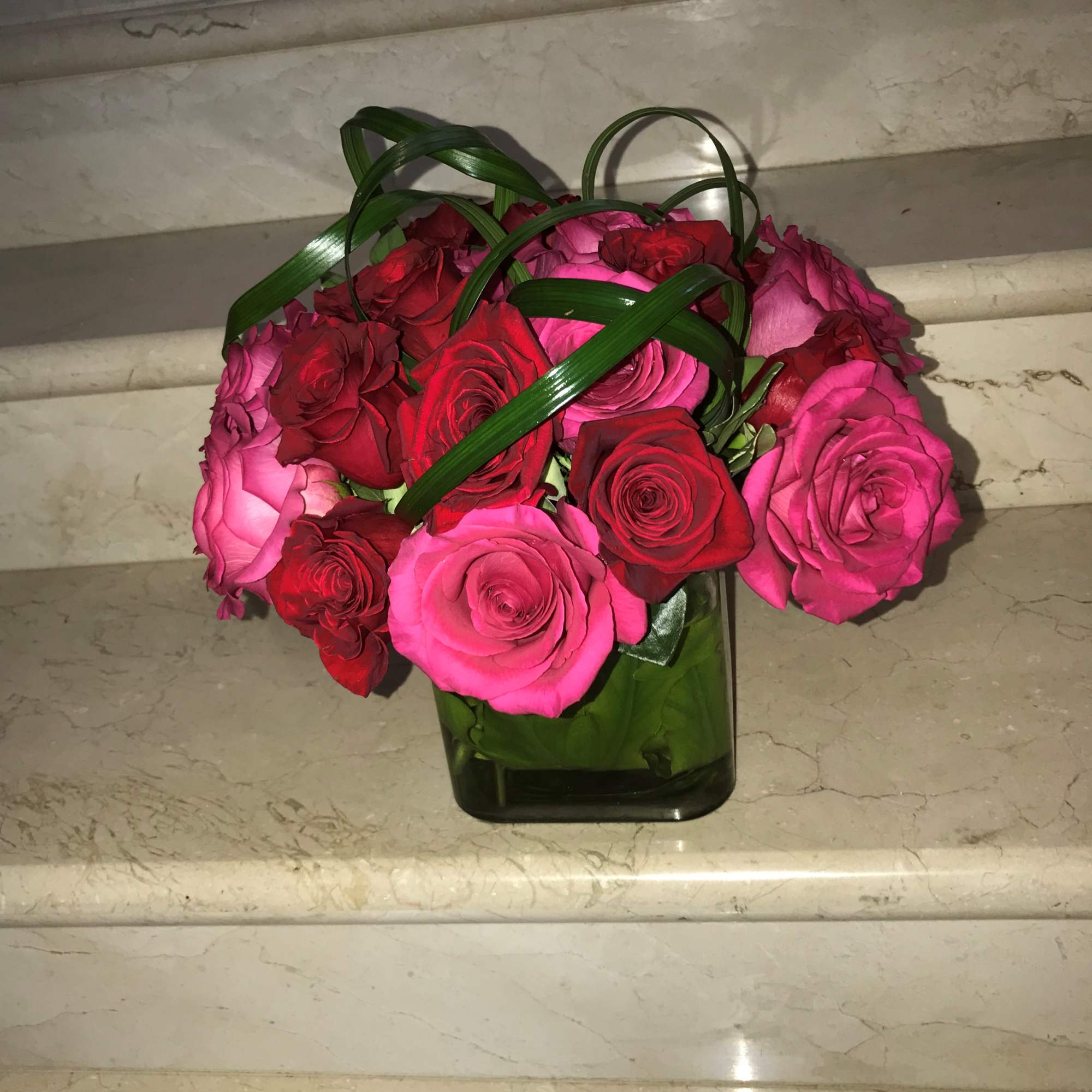 Compact arrangement of red and hot pink roses in a square glass vase with looped green blades.