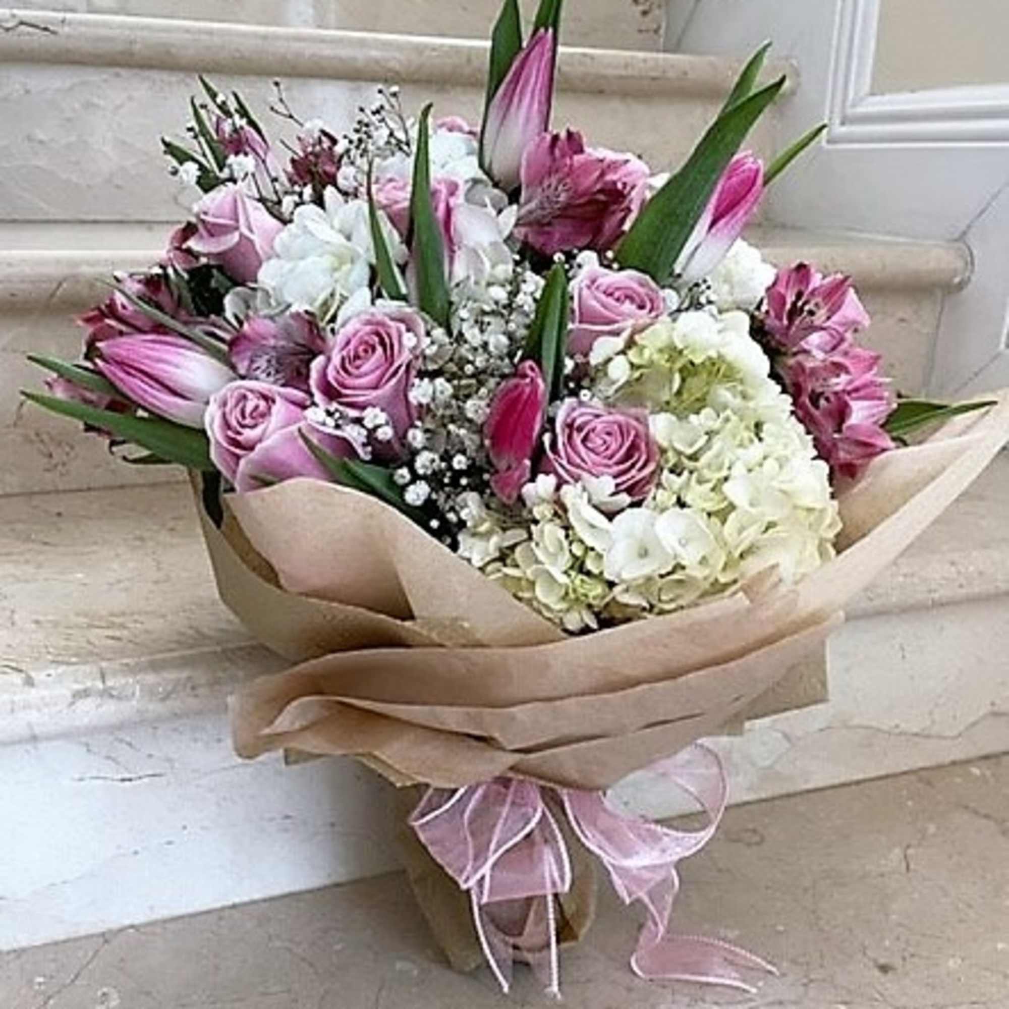 Hand-tied bouquet of pink roses, white hydrangeas, and pink lilies wrapped in kraft paper with a pink ribbon