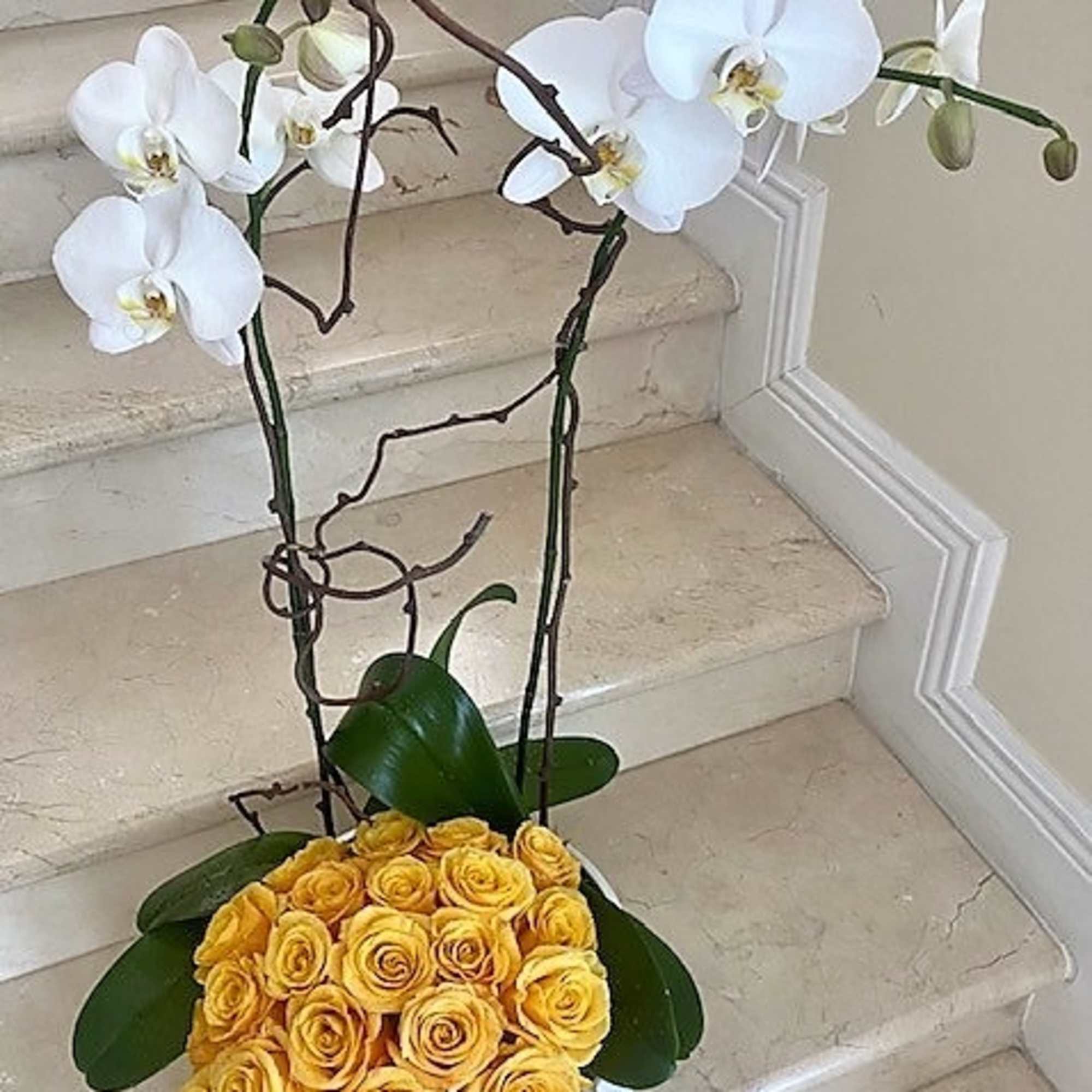 Tall arrangement with white orchids on branches above a dome of yellow roses in a white bowl