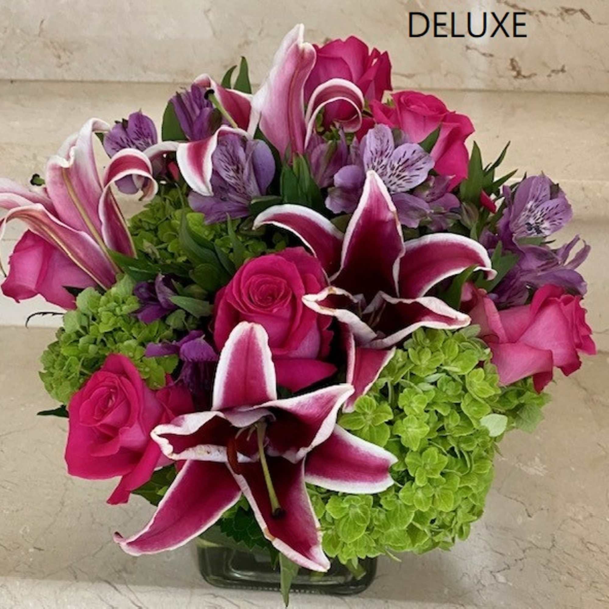 Low cube arrangement of hot pink roses, pink lilies, purple blooms, and green hydrangeas in a glass vase