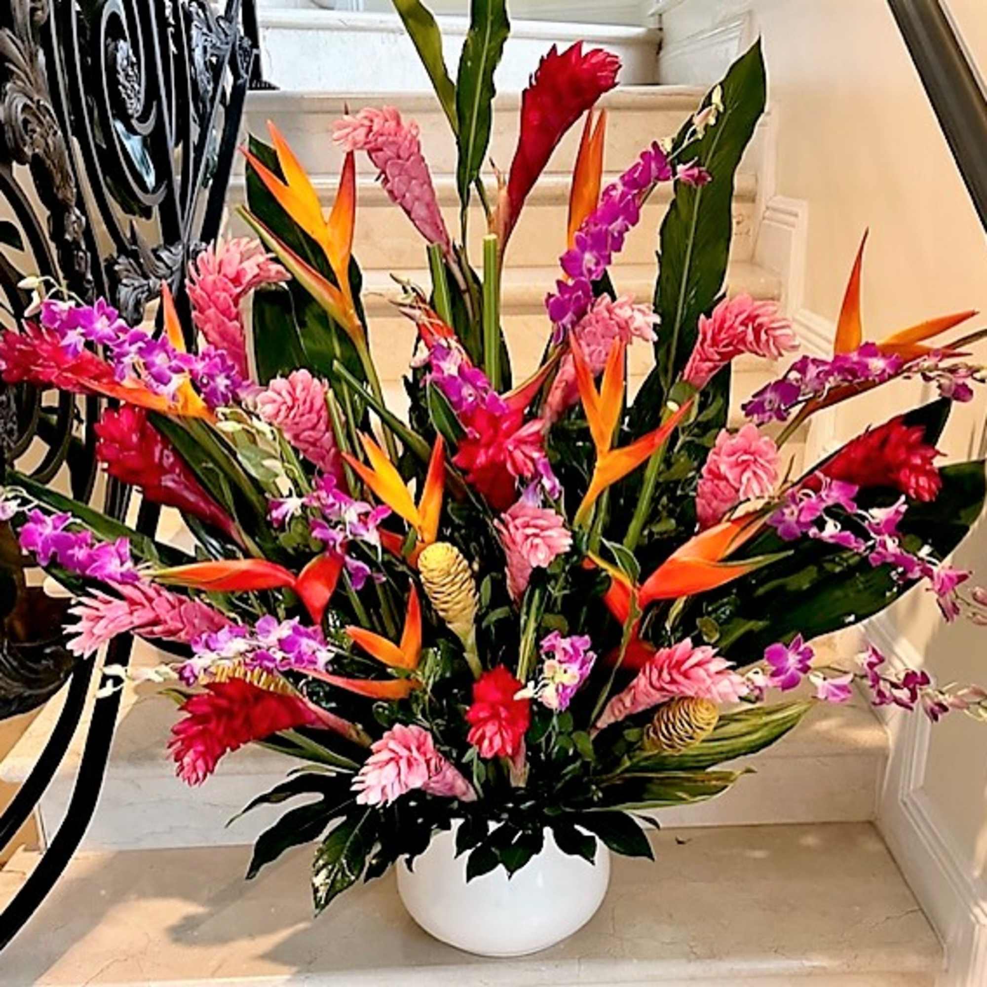 Large tropical arrangement with pink gingers, orange birds of paradise and purple orchids in a white bowl vase