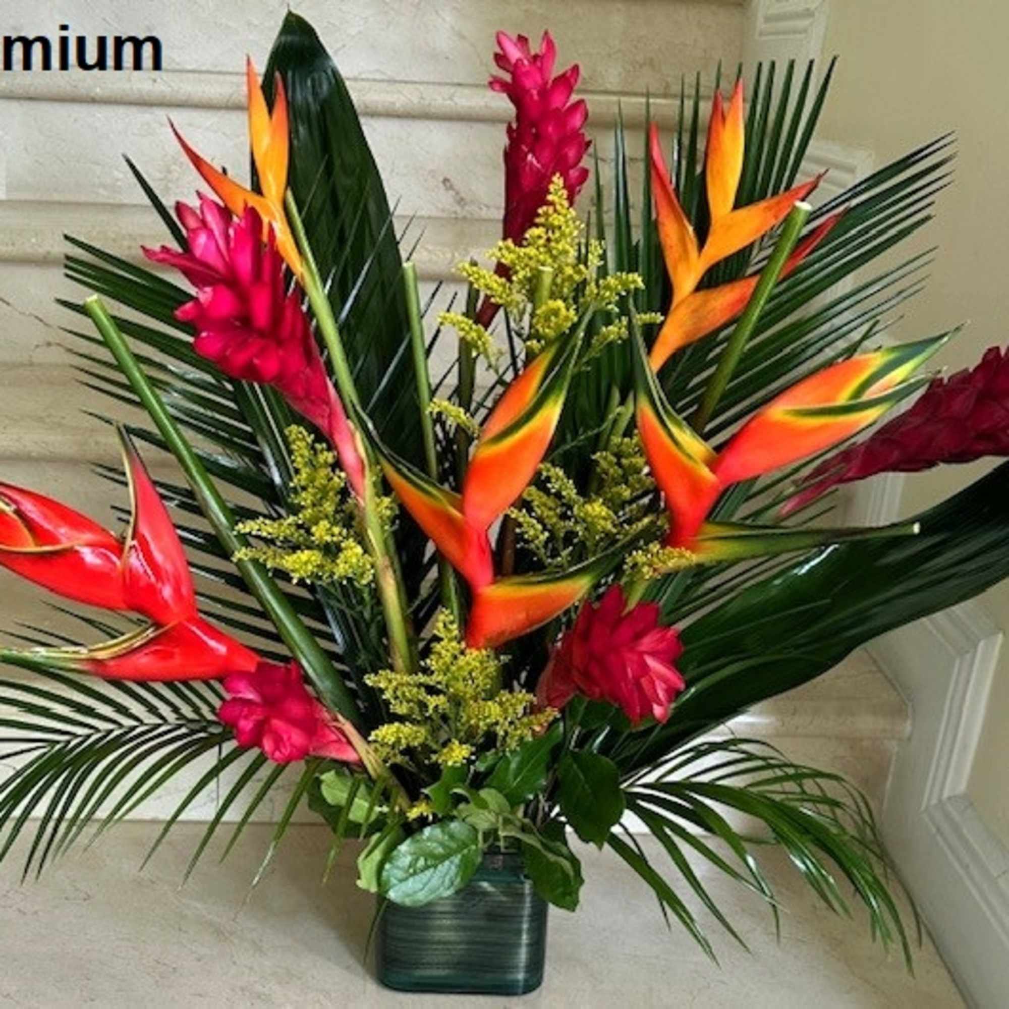 Large tropical arrangement with orange and red flowers and tall green leaves in a square vase