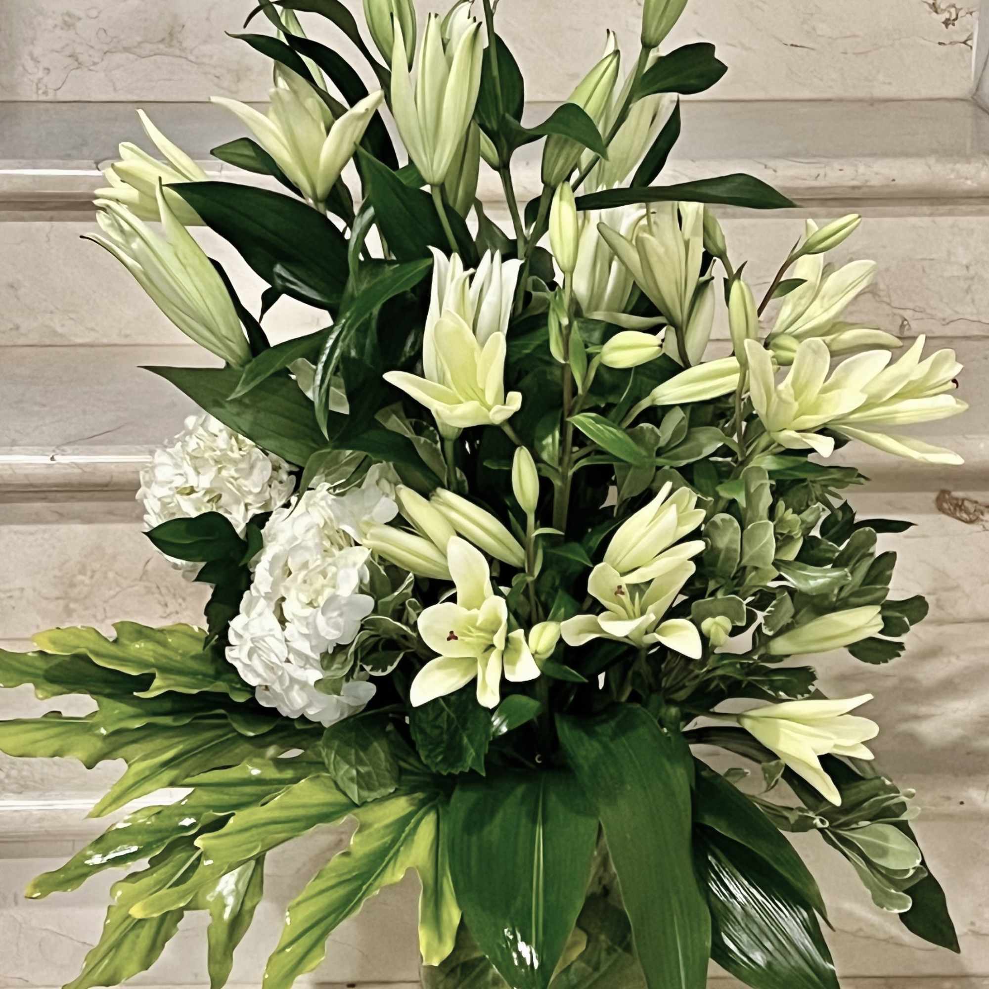 Tall arrangement of white lilies and hydrangeas with bold foliage in a green glass vase