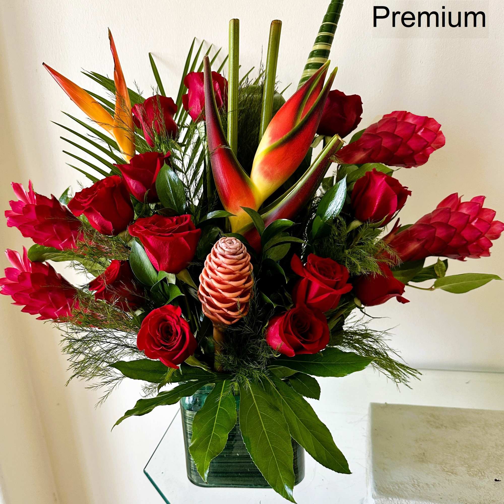 Tall tropical arrangement with red roses and exotic flowers in a green glass cube vase