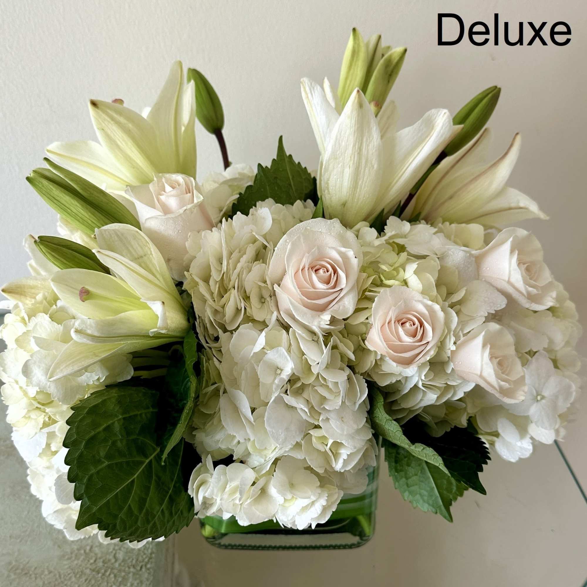 Compact arrangement of white lilies, hydrangeas, and pale roses in a clear glass cube vase.