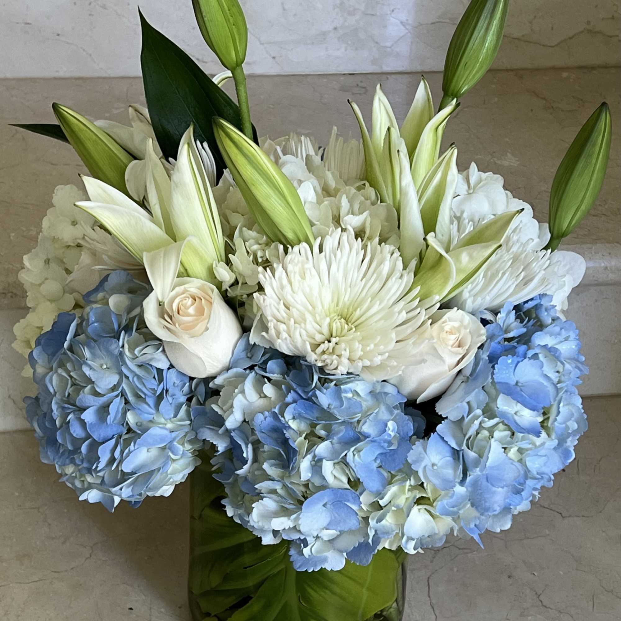 Arrangement of blue hydrangeas, white lilies, roses, and mums in a clear glass vase wrapped with green leaves.