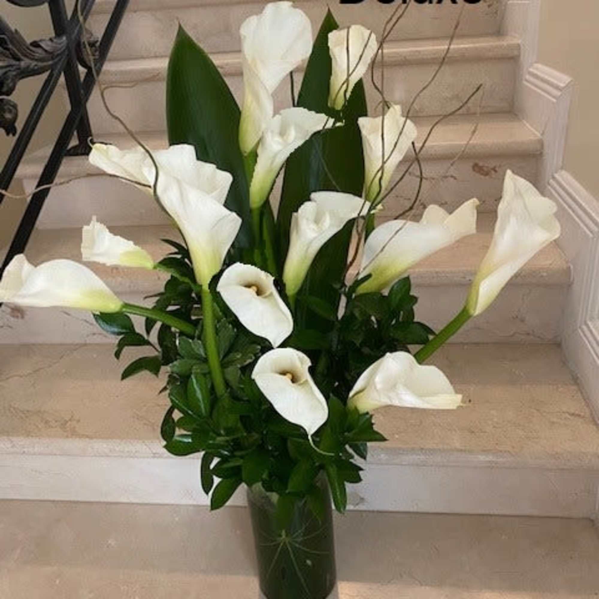 Tall arrangement of white calla lilies in a green-filled glass vase on stone steps