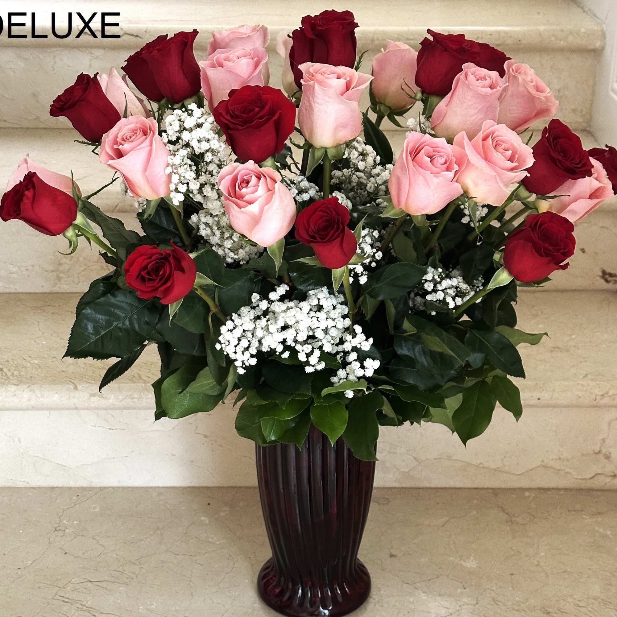 Tall arrangement of red and pink roses with white filler flowers in a dark red glass vase