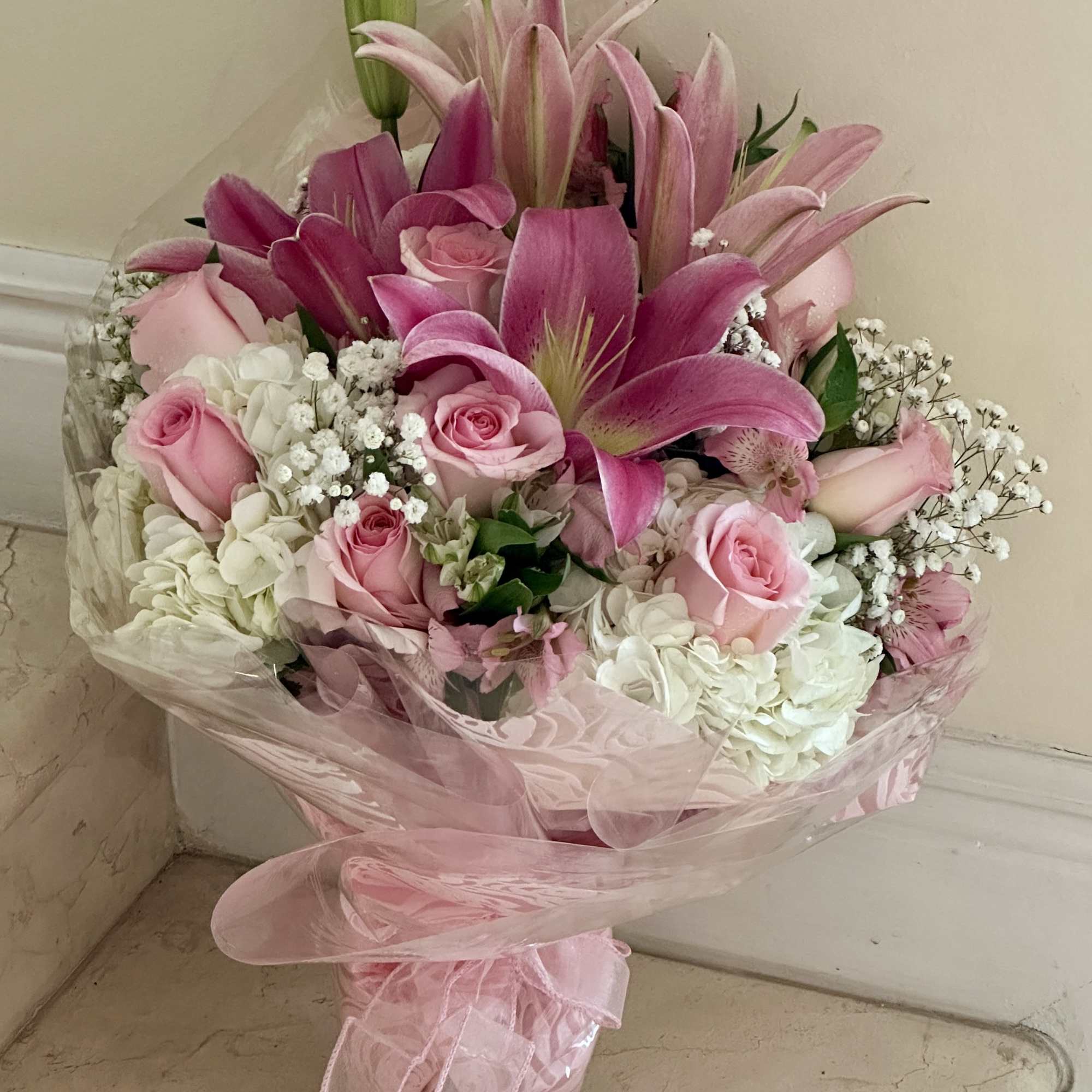 Hand-tied bouquet of pink lilies and roses with white hydrangeas wrapped in pink cellophane.