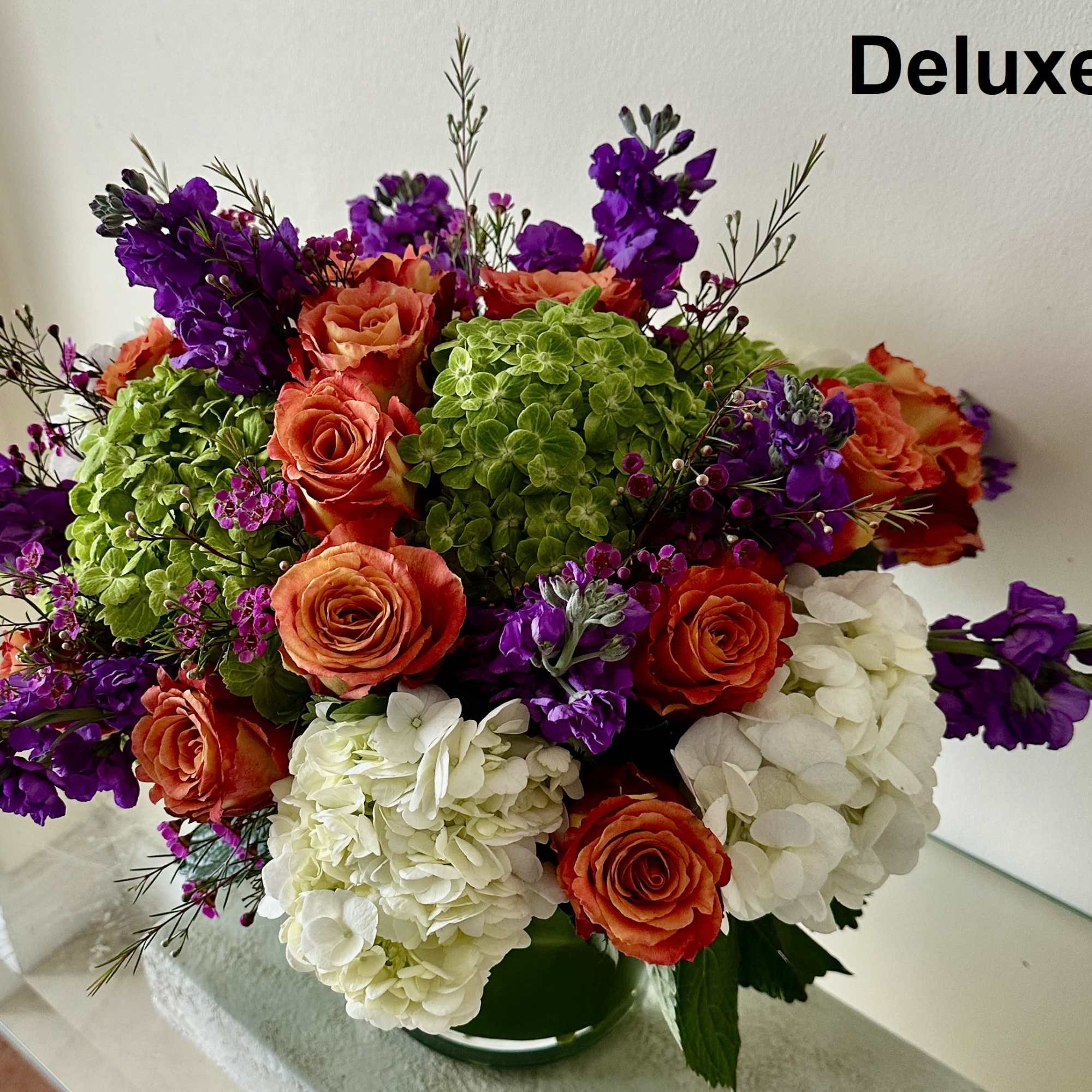 Round arrangement of orange roses, green and white hydrangeas, and purple stock in a low glass vase