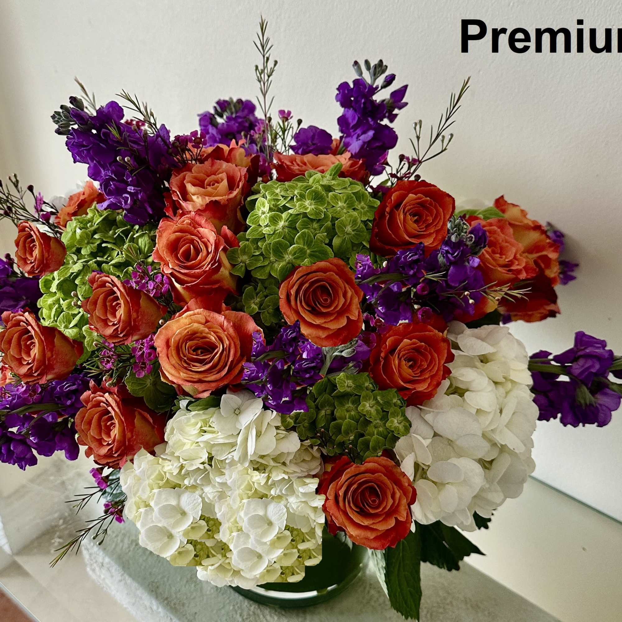 Low vase arrangement of orange roses, purple stock, and green and white hydrangeas