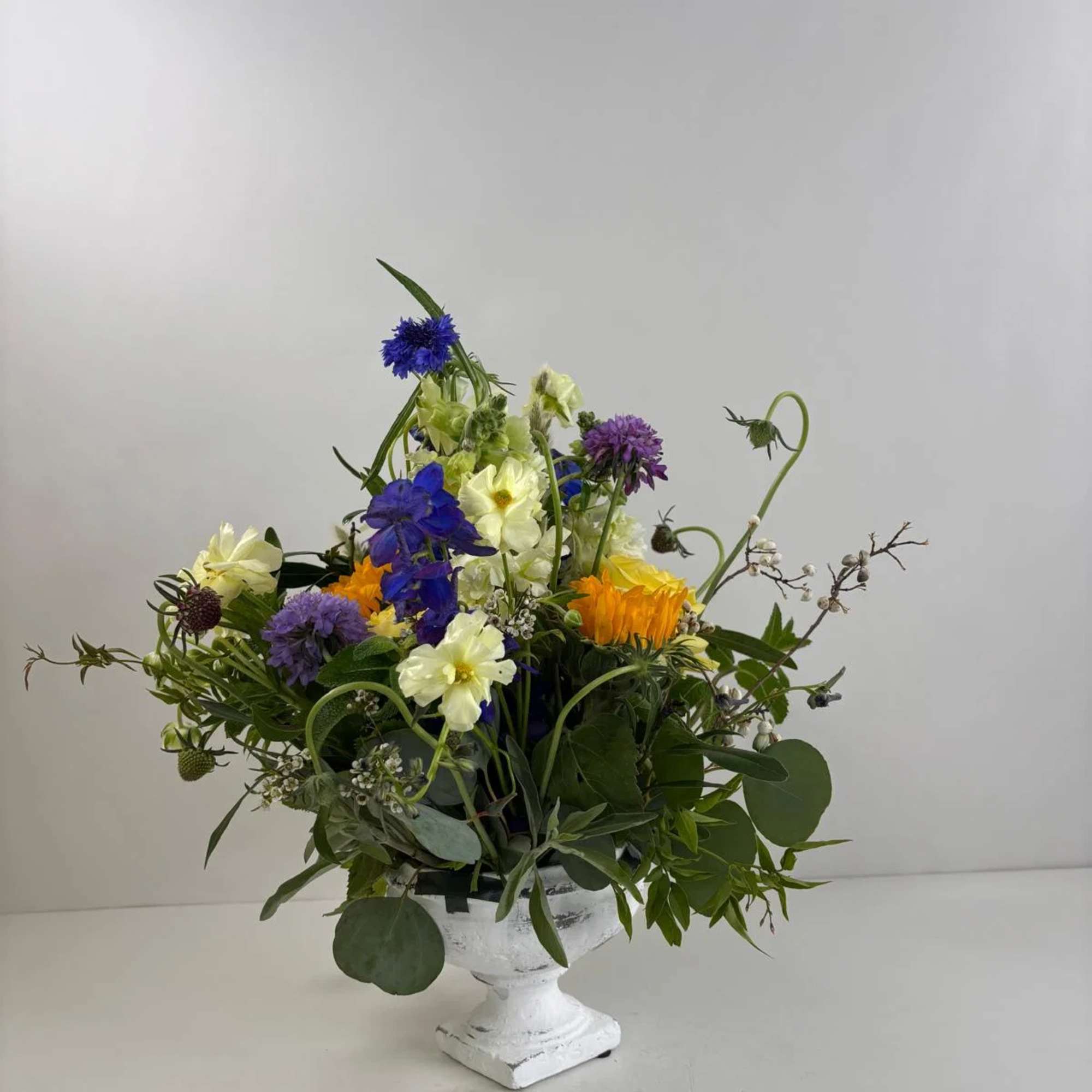 Mixed bouquet in a white pedestal vase with blue, yellow, purple, and orange flowers