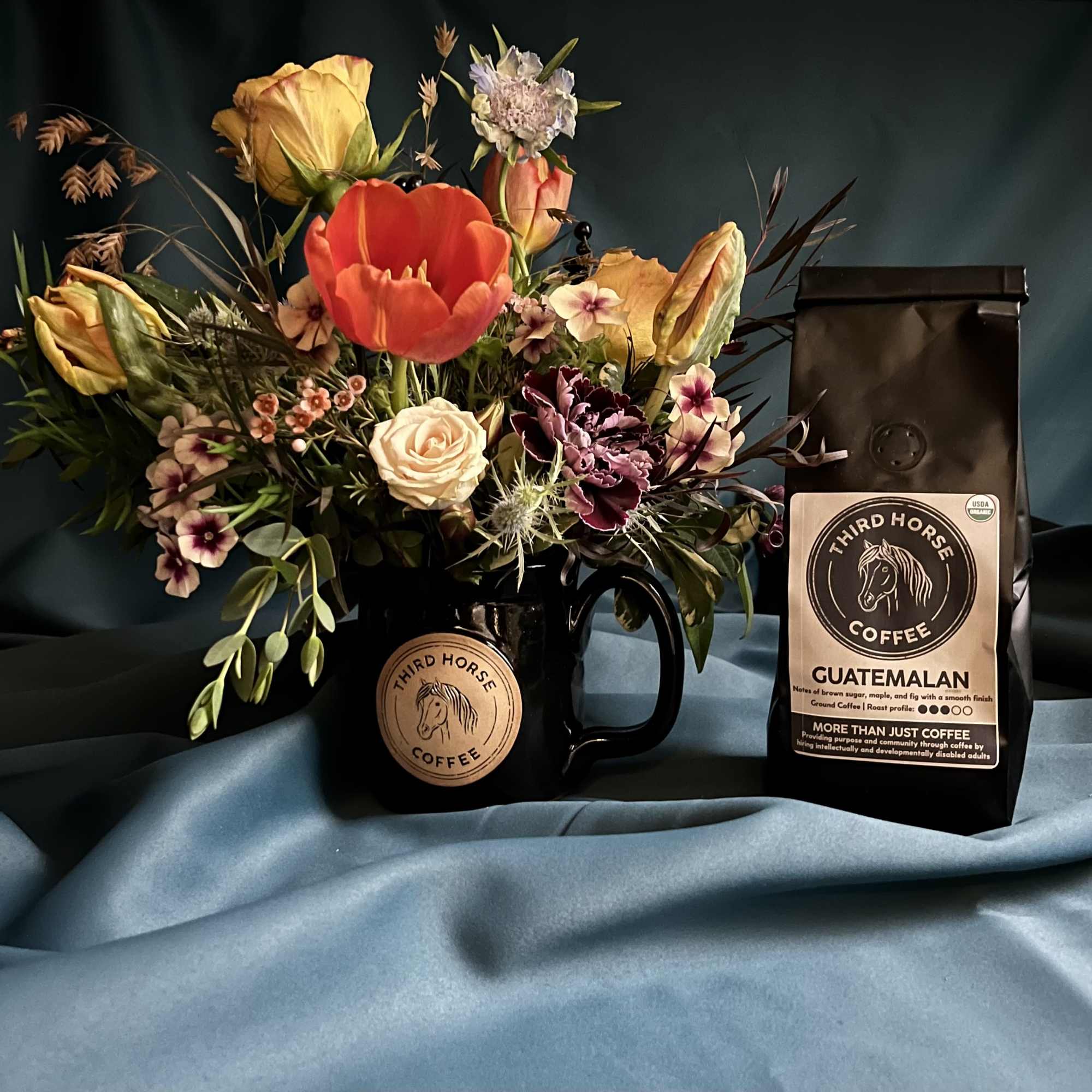 Mug of mixed flowers beside a branded bag of Guatemalan coffee on teal fabric.
