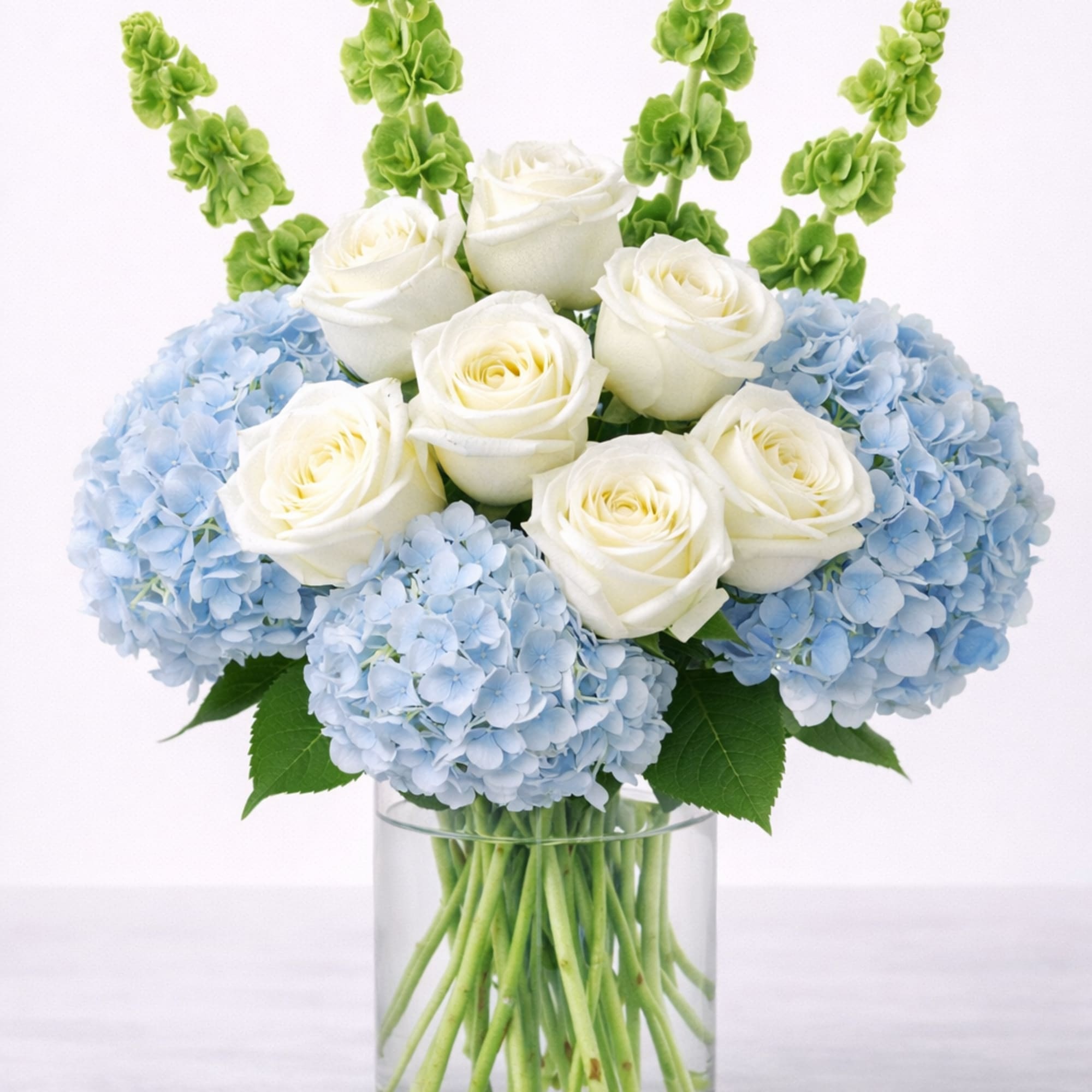 White roses and blue hydrangeas in a clear glass vase with green spikes
