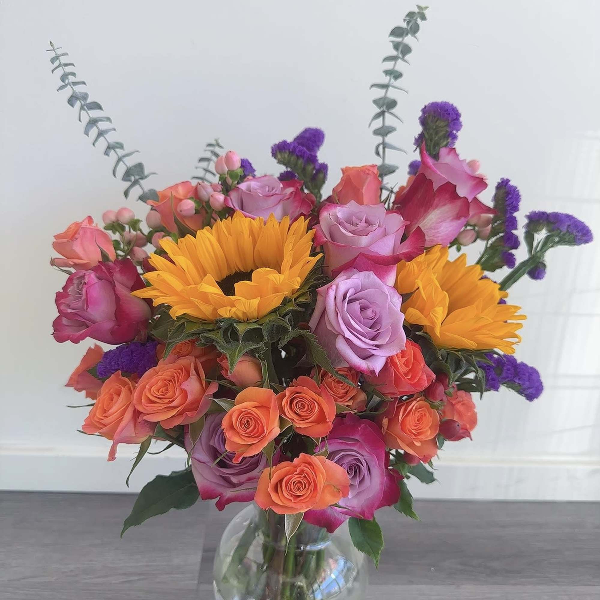 Mixed bouquet with sunflowers, roses, and purple filler in a glass vase