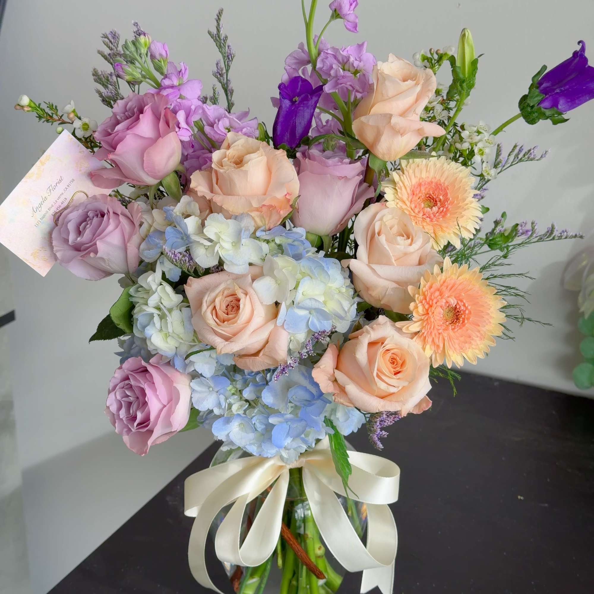 Mixed pastel bouquet of roses, hydrangeas, and purple blooms in a glass vase with ribbon