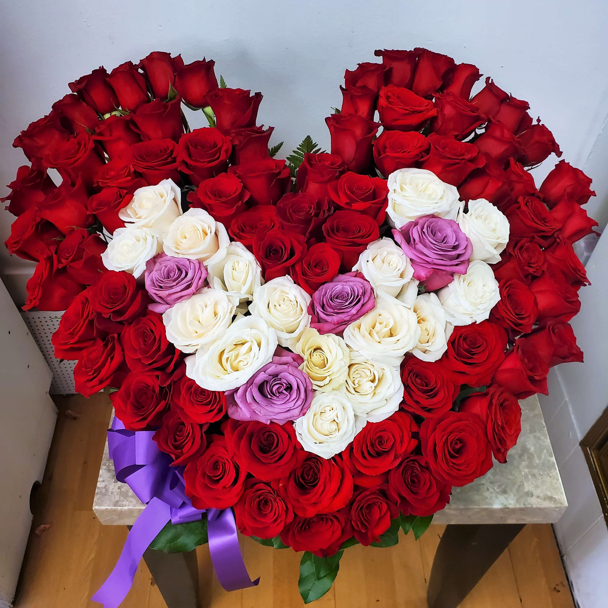 Heart-shaped bouquet of red, white, and lavender roses with a purple ribbon
