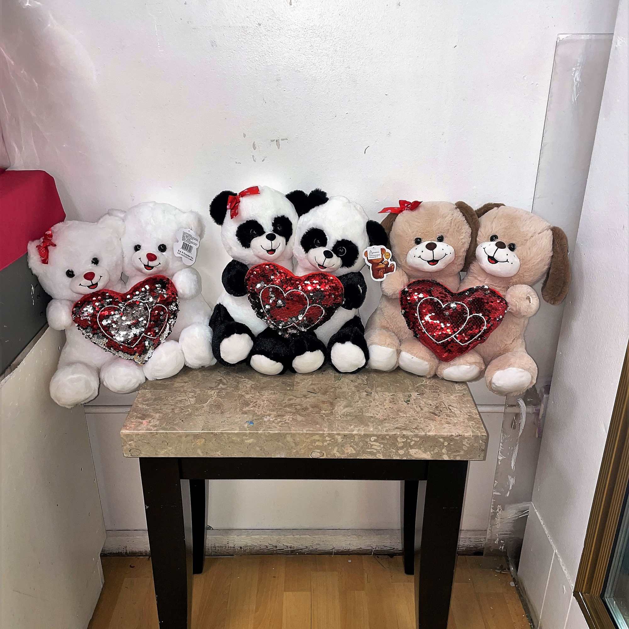 Three plush teddy bears and two panda plush toys holding red heart pillows on a table.