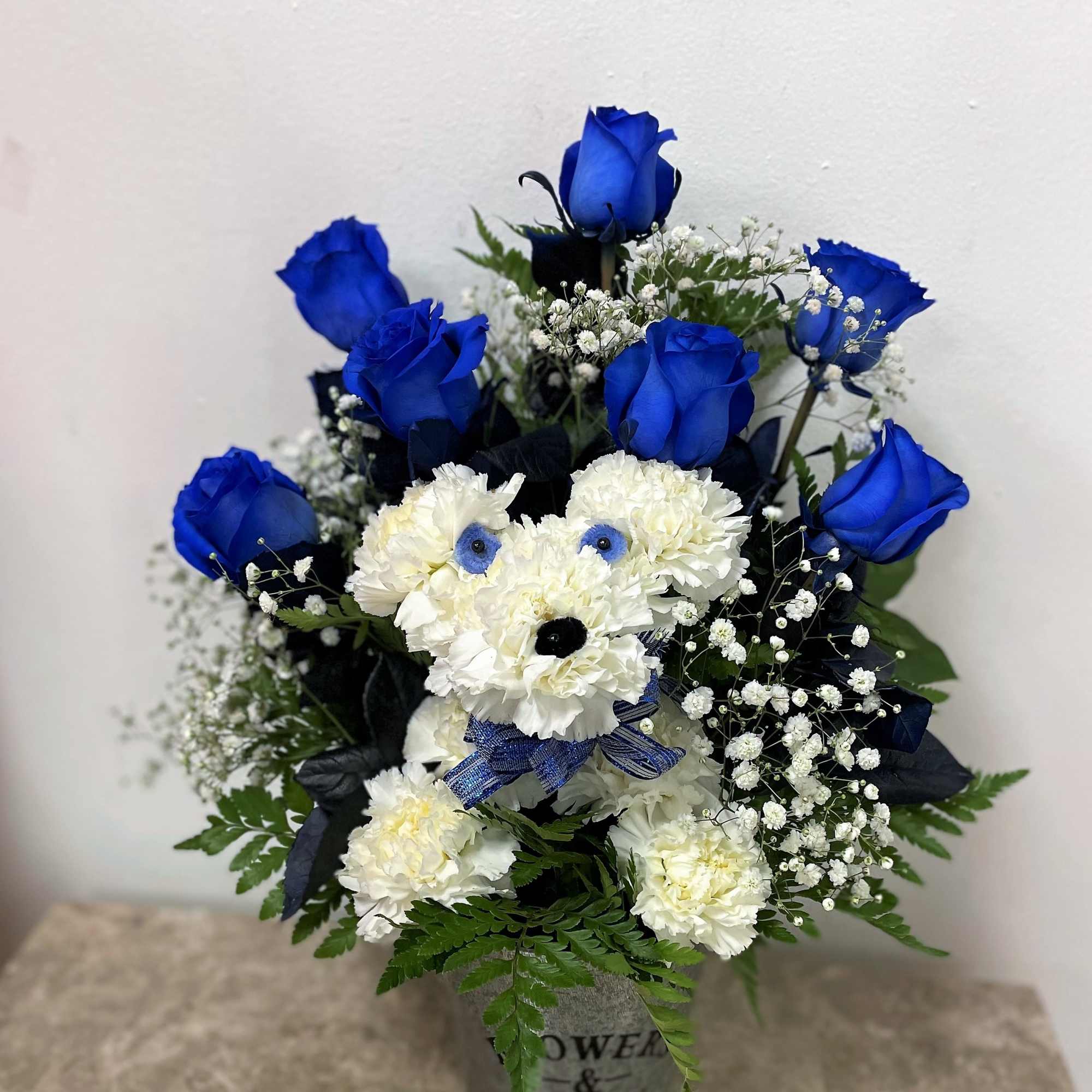 Blue roses and white flowers arranged in a vase with a teddy bear face.