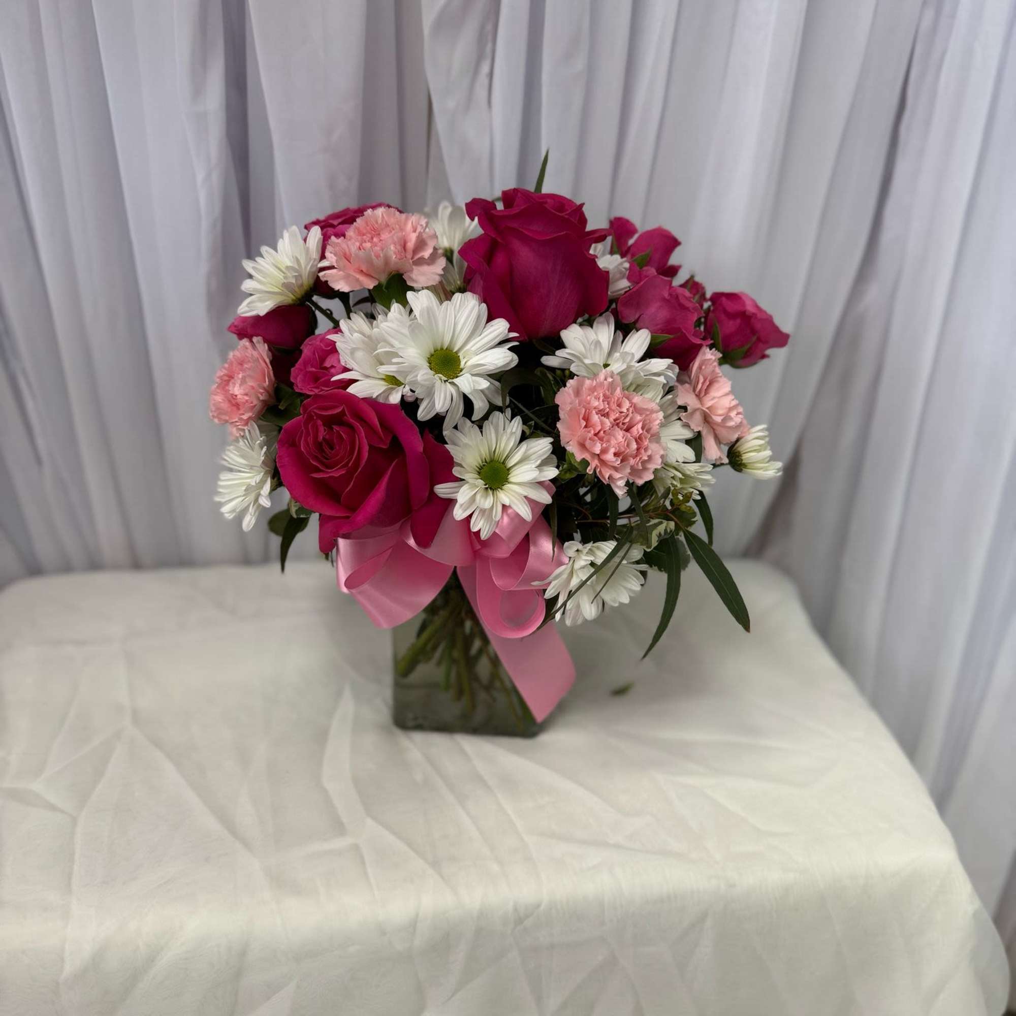 Pink roses and white daisies arranged in a glass vase with a pink ribbon