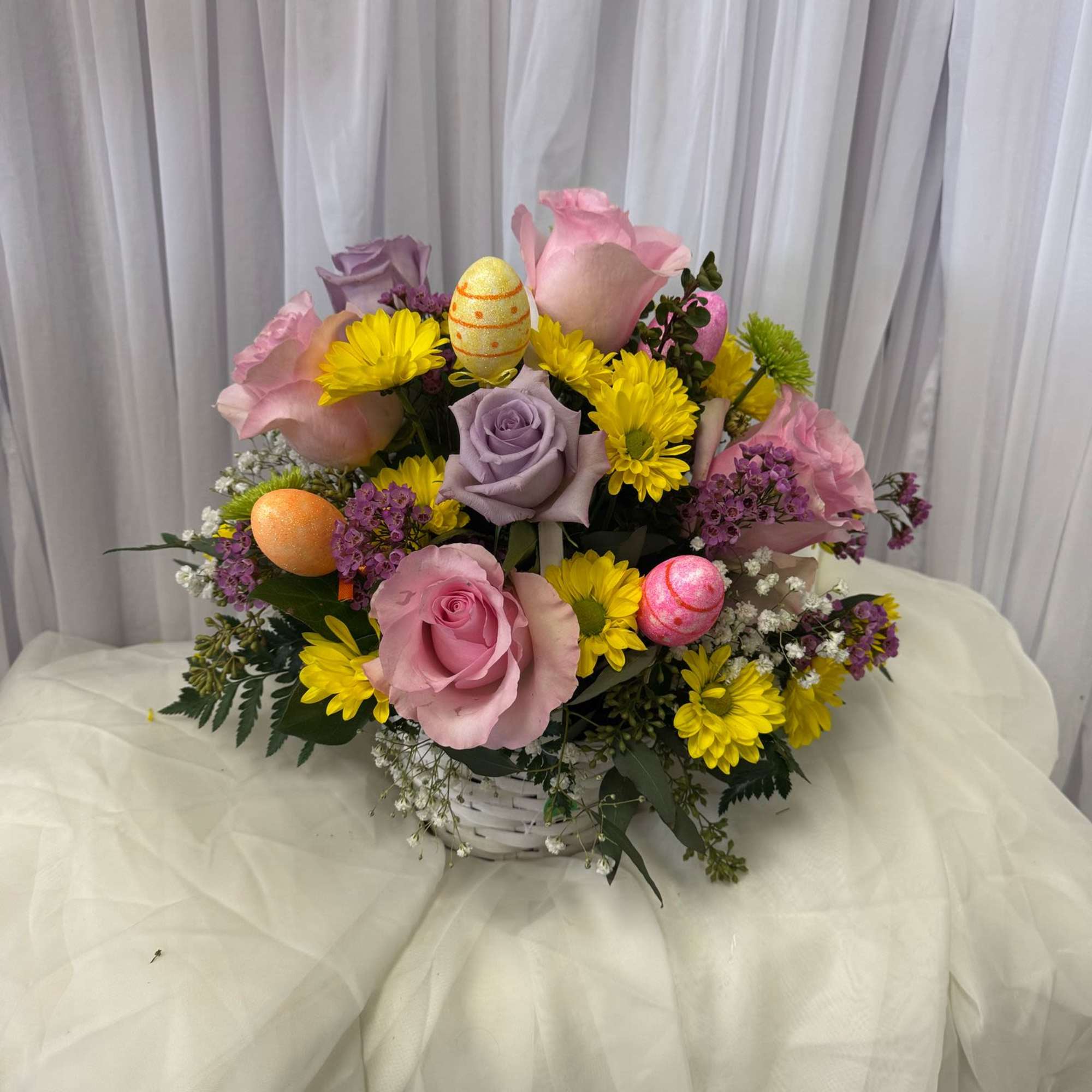 Basket arrangement of pink and lavender roses with yellow daisies and Easter eggs