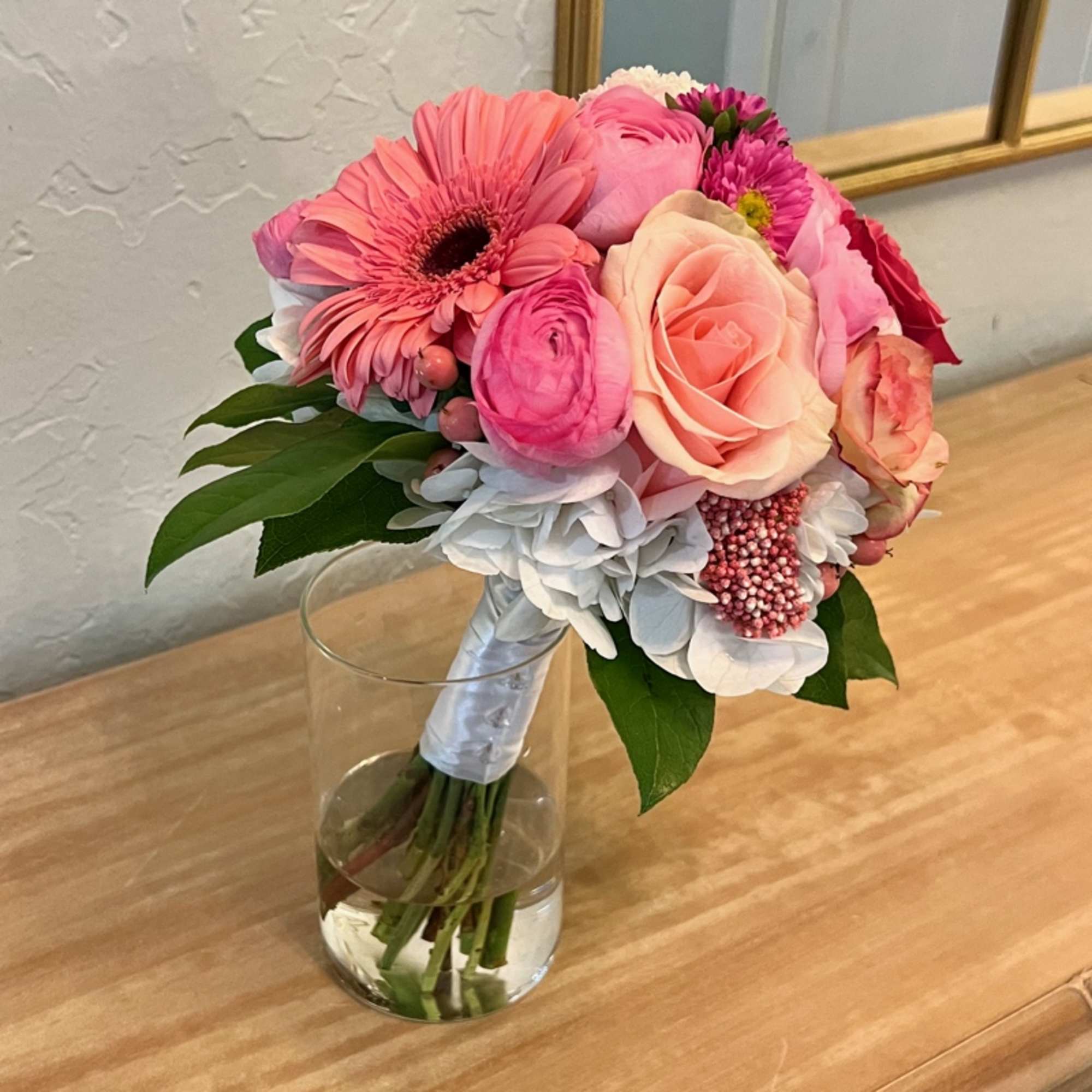 Compact bouquet of pink gerbera daisies, roses, and ranunculus with white hydrangea in a clear glass vase.
