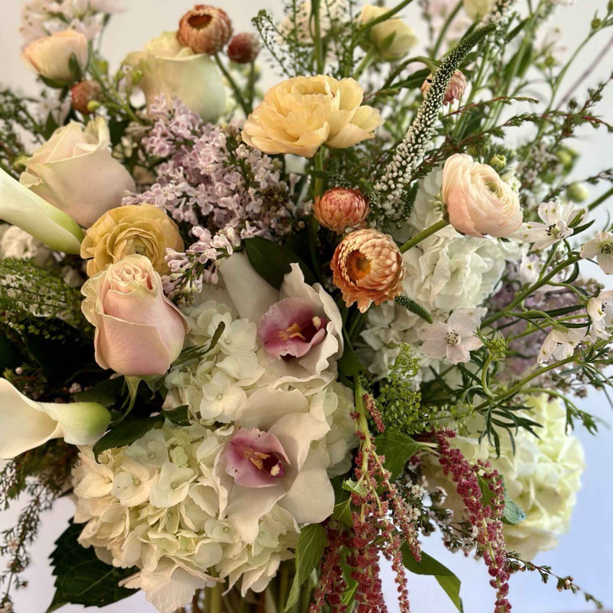 Tall mixed arrangement of white hydrangeas, pale roses, ranunculus, and calla lilies in a glass vase
