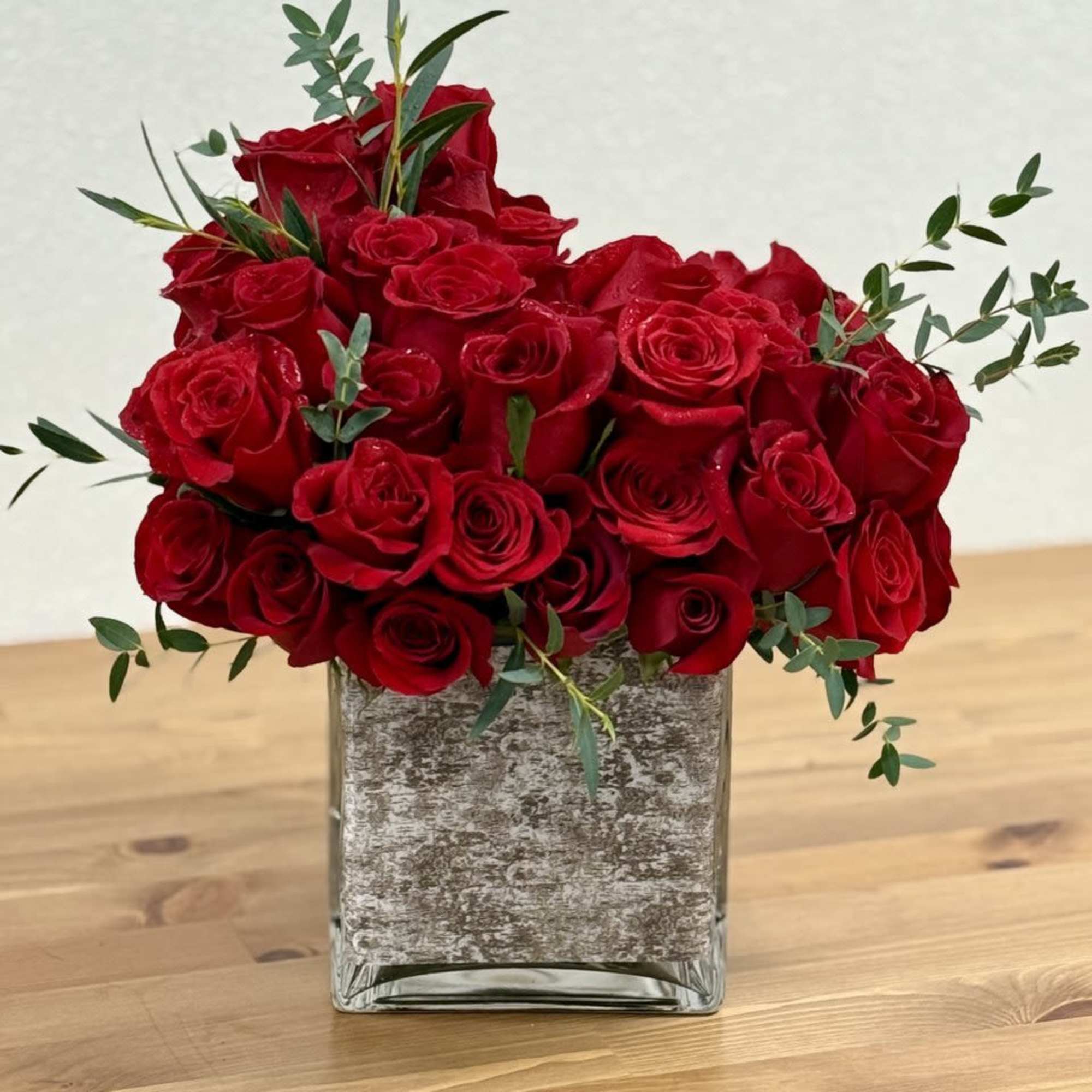 Red roses arranged in a square glass vase with sparse greenery