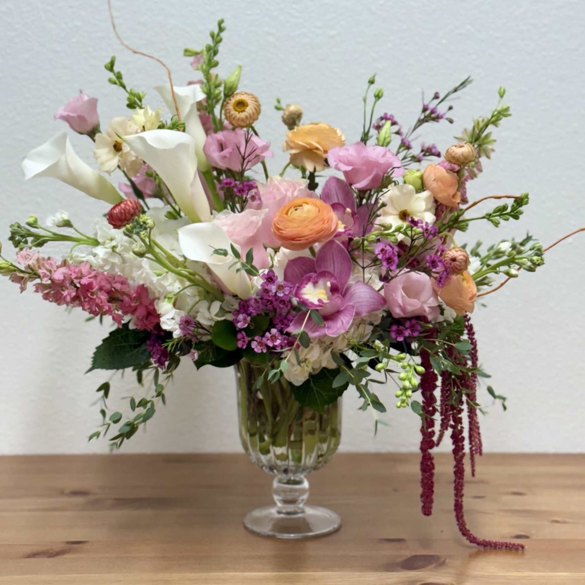 Pastel arrangement of pink, peach, and white flowers with calla lilies in a clear footed glass vase