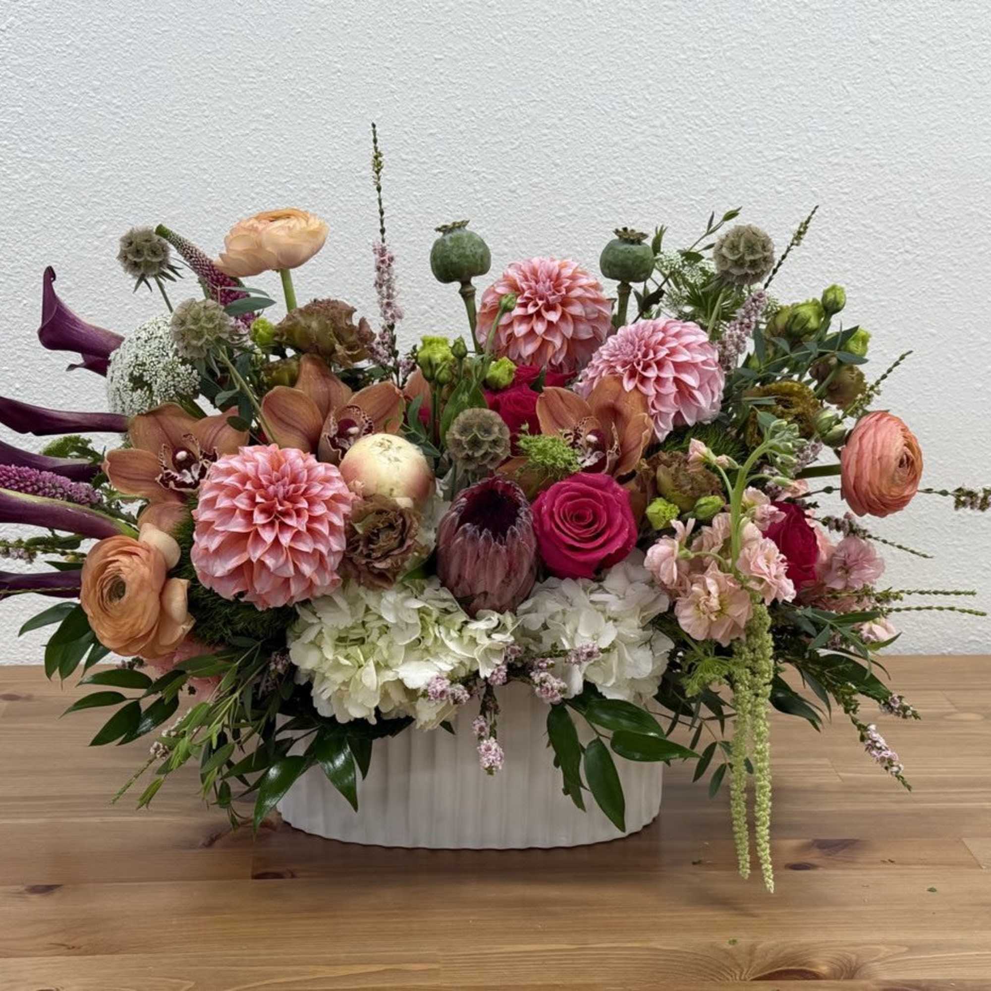 Low oval arrangement of pink dahlias, roses, calla lilies and mixed blooms in a white ceramic vase.