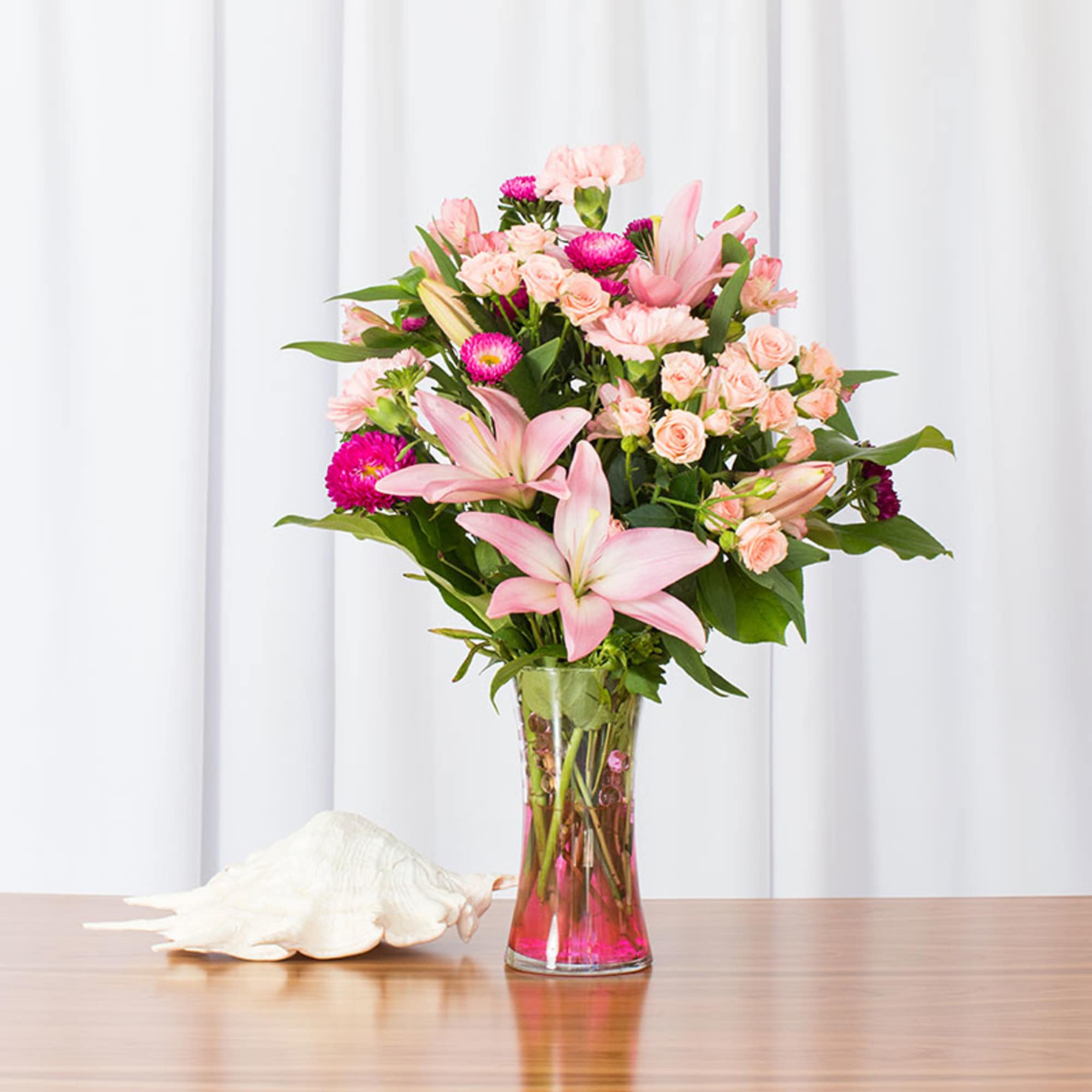 Tall glass vase of pink lilies, peach roses, and pink flowers on a table beside a large white seashell