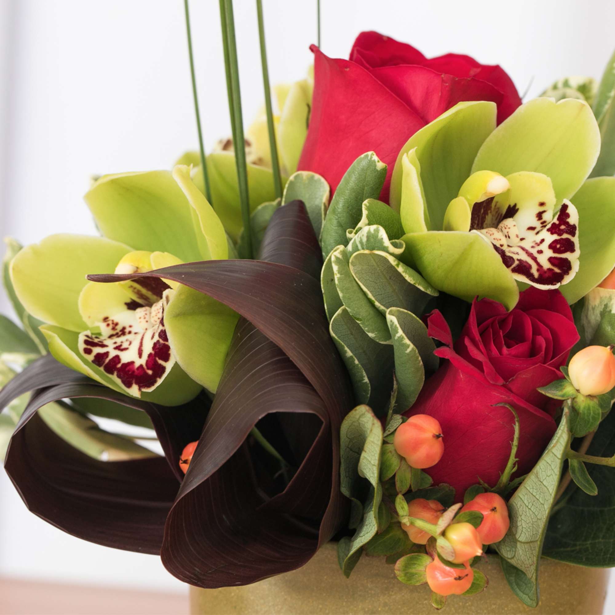Compact arrangement of red roses and green orchids with dark foliage in a gold vase