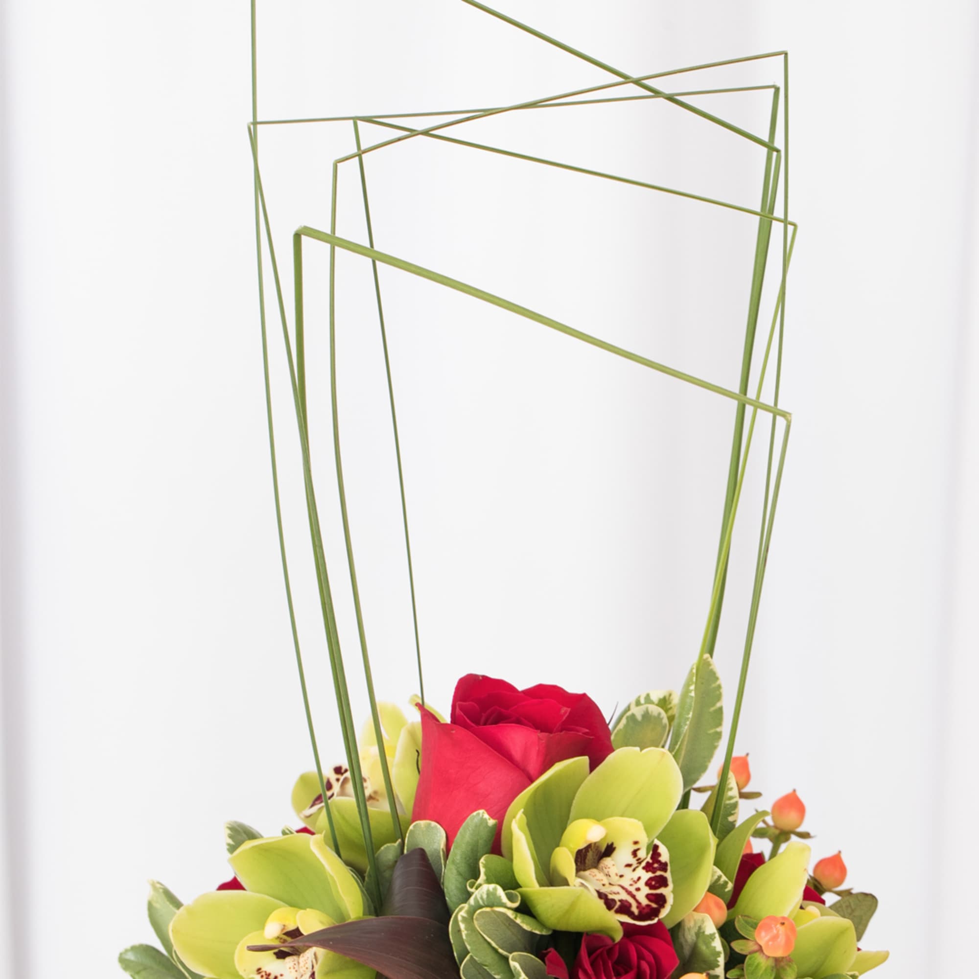 Modern arrangement of red roses and green cymbidium orchids with tall geometric grass loops
