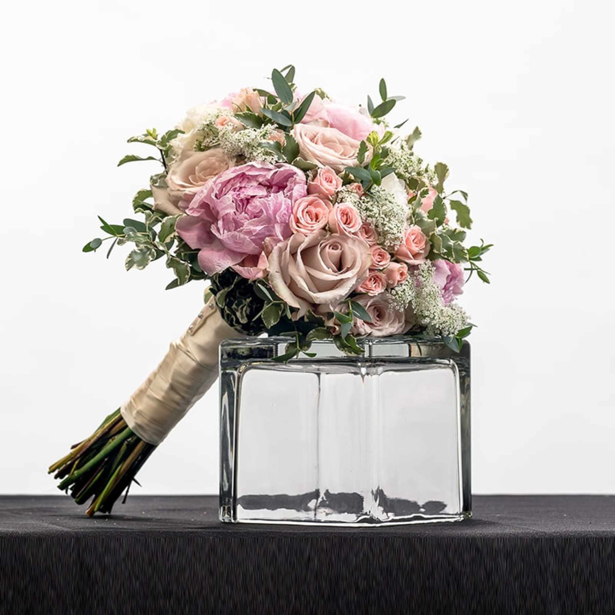 Hand-tied bouquet of blush roses and pink peonies resting on a clear glass block vase