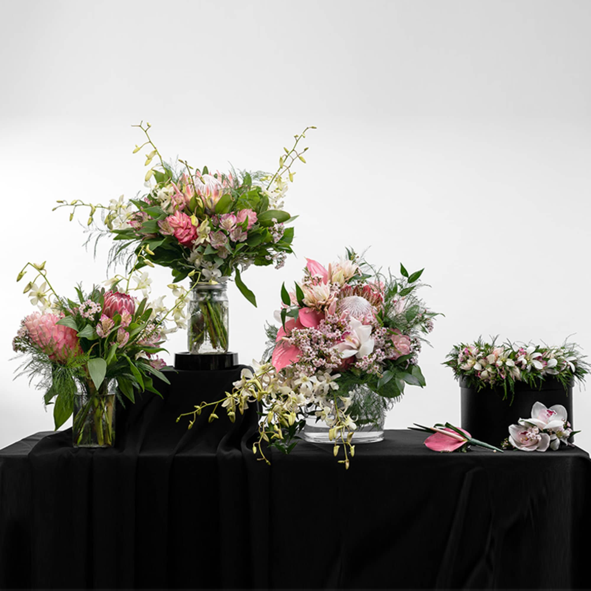 Group of pink and white protea and orchid arrangements in clear vases and a black hat box on a black tablecloth.