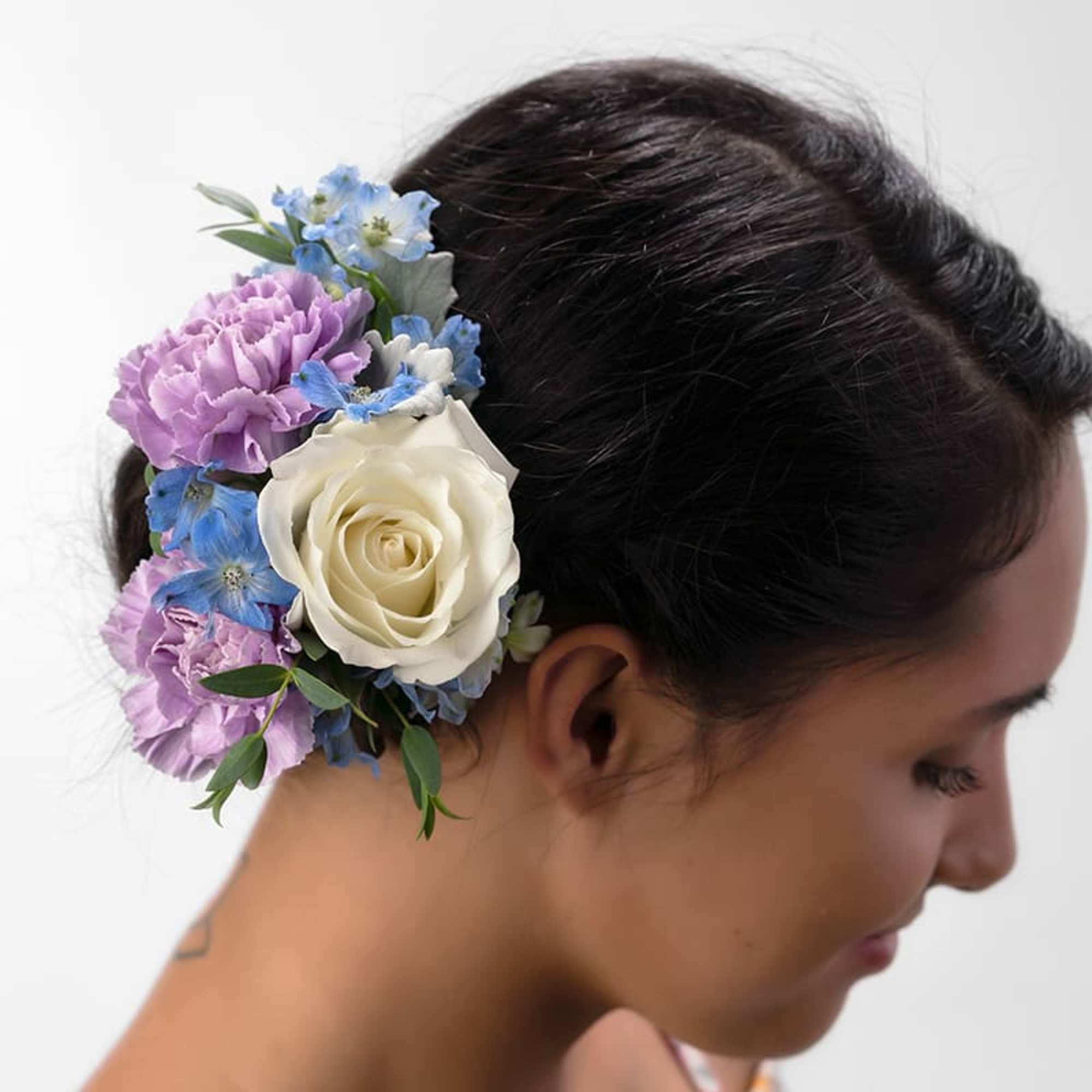 Floral hairpiece with white rose, purple carnations, and blue flowers worn in dark hair.