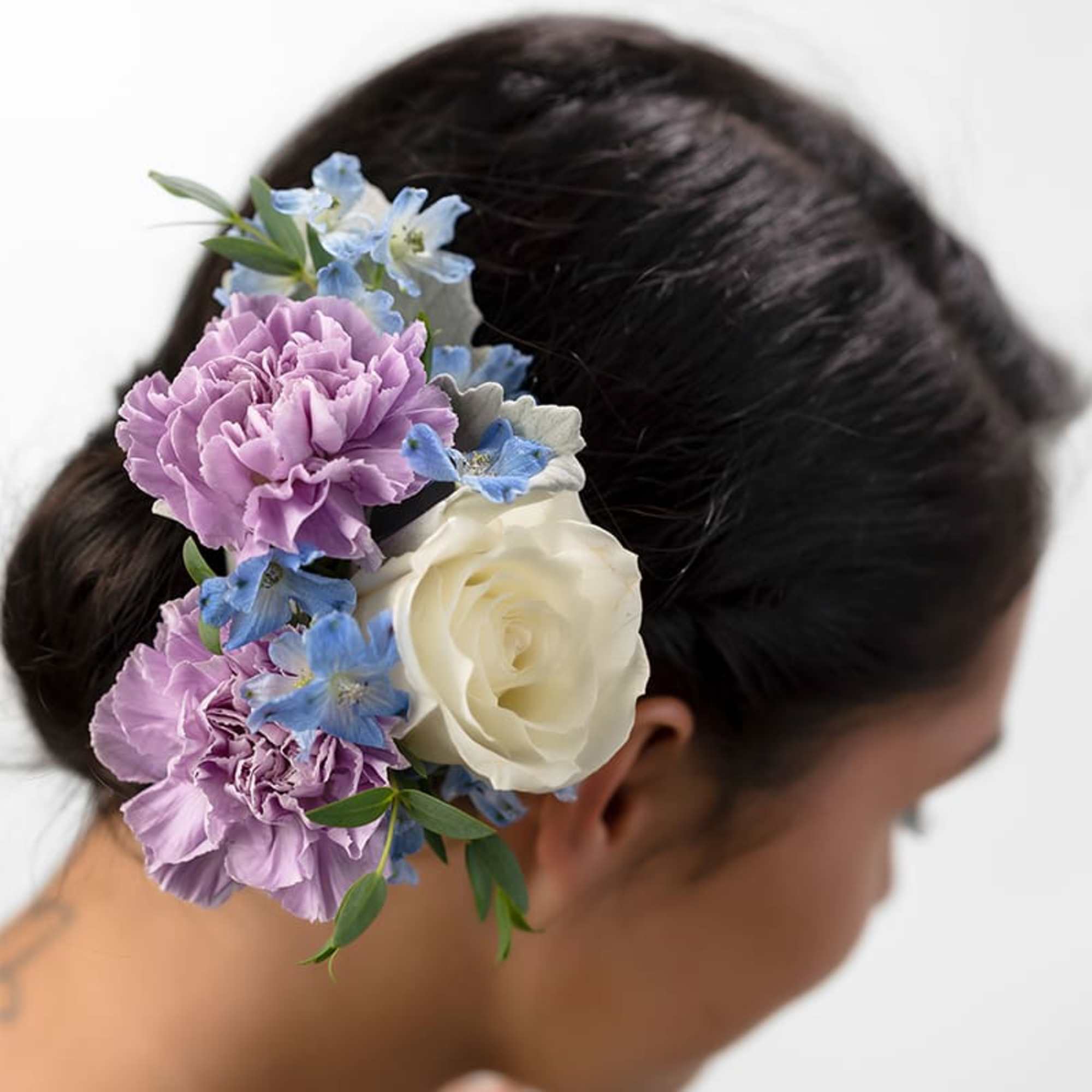 Floral hairpiece with lavender carnations, a white rose, and blue blooms worn in a dark updo.