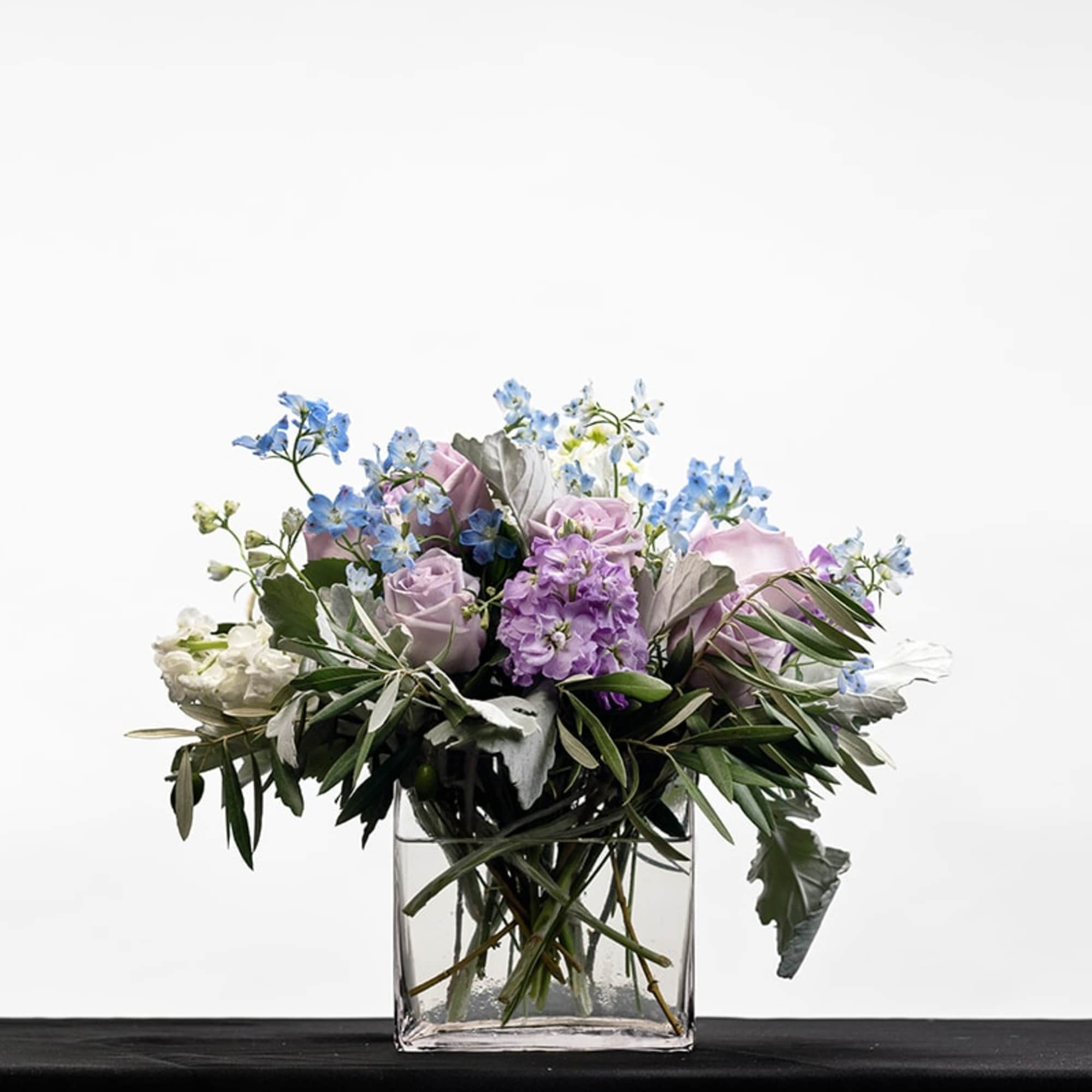 Lavender roses, purple blooms, and blue flowers arranged in a clear glass cube vase